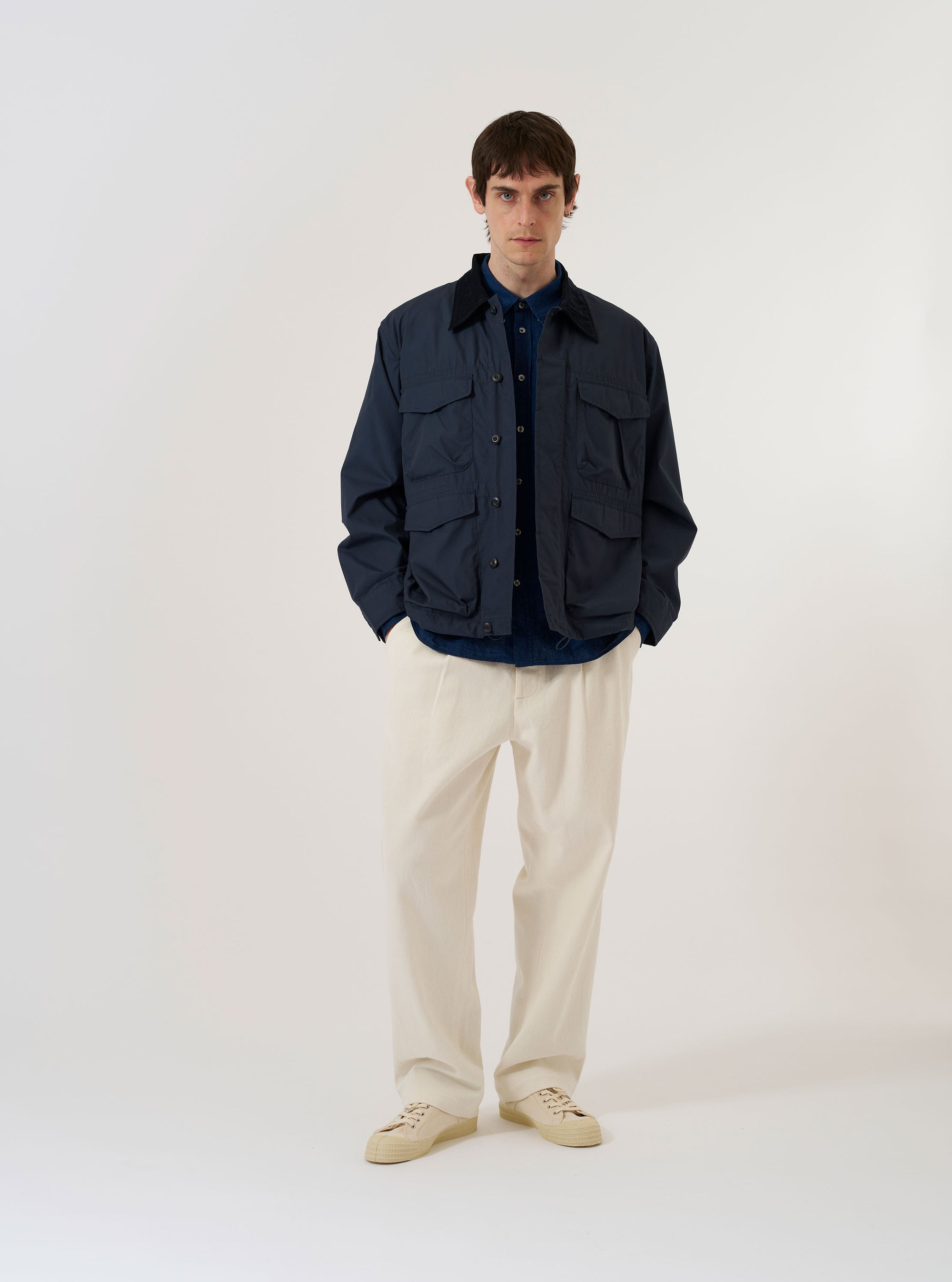 A model wears the Navy Recycled Polytech Parachute Field Jacket over a light-colored shirt and olive trousers, standing against a plain white background. The jacket features a contrasting brown corduroy collar, a buttoned neck strap, and four large 3D patch pockets with button flaps on the front. The garment has a loose, utilitarian fit with a straight hem and adjustable button cuffs.