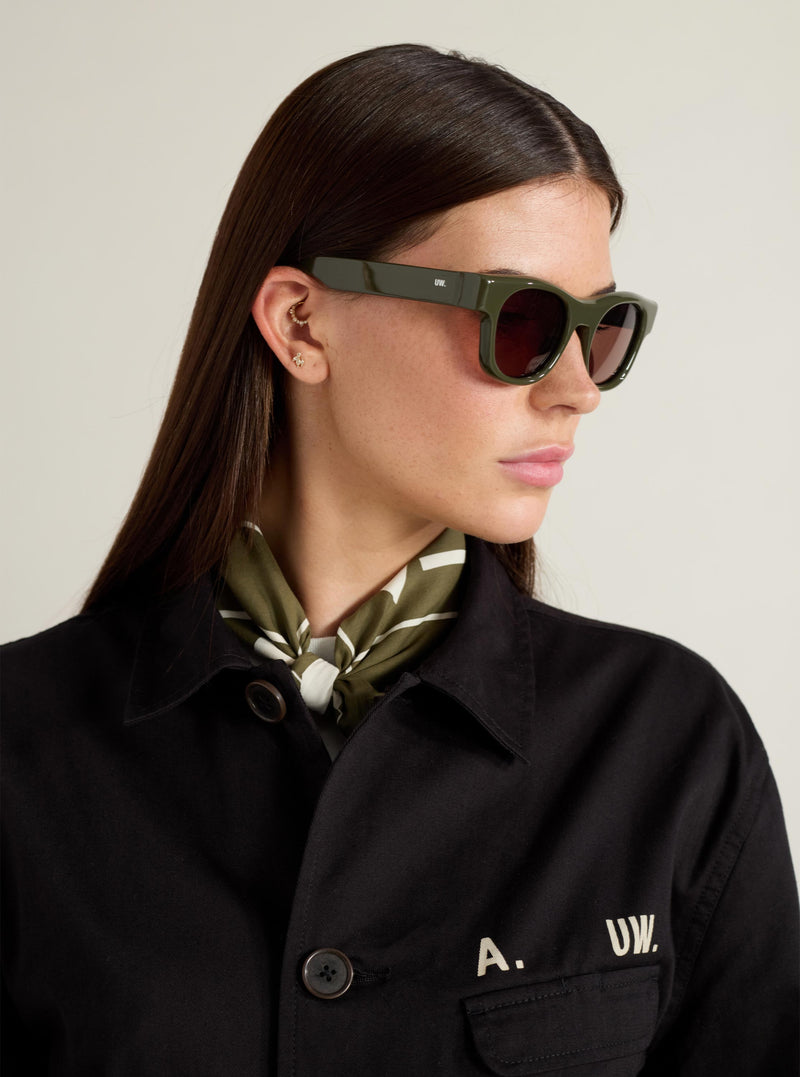 Model wearing A. SOCIETY x Universal Works Olive Stanley Sunglasses. The olive frames complement her black jacket and patterned scarf, set against a neutral background.