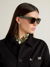 Model wearing A. SOCIETY x Universal Works Olive Stanley Sunglasses. The olive frames complement her black jacket and patterned scarf, set against a neutral background.