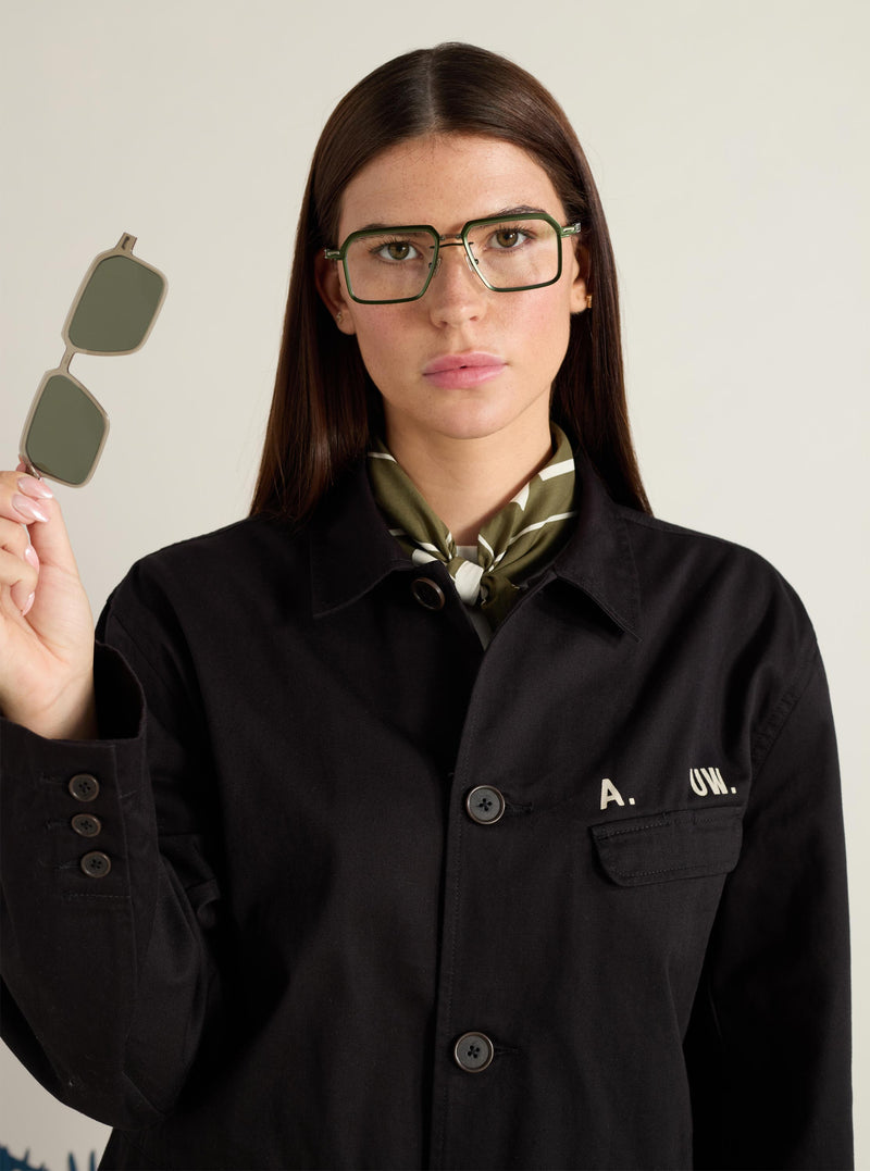 Model presents A. SOCIETY x Universal Works Olive/Green Kowloon Eyewear. She holds the olive clip-on lenses. The glasses have a green and gold metal frame. Studio shot on a neutral background.