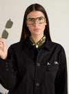 Model presents A. SOCIETY x Universal Works Olive/Green Kowloon Eyewear. She holds the olive clip-on lenses. The glasses have a green and gold metal frame. Studio shot on a neutral background.