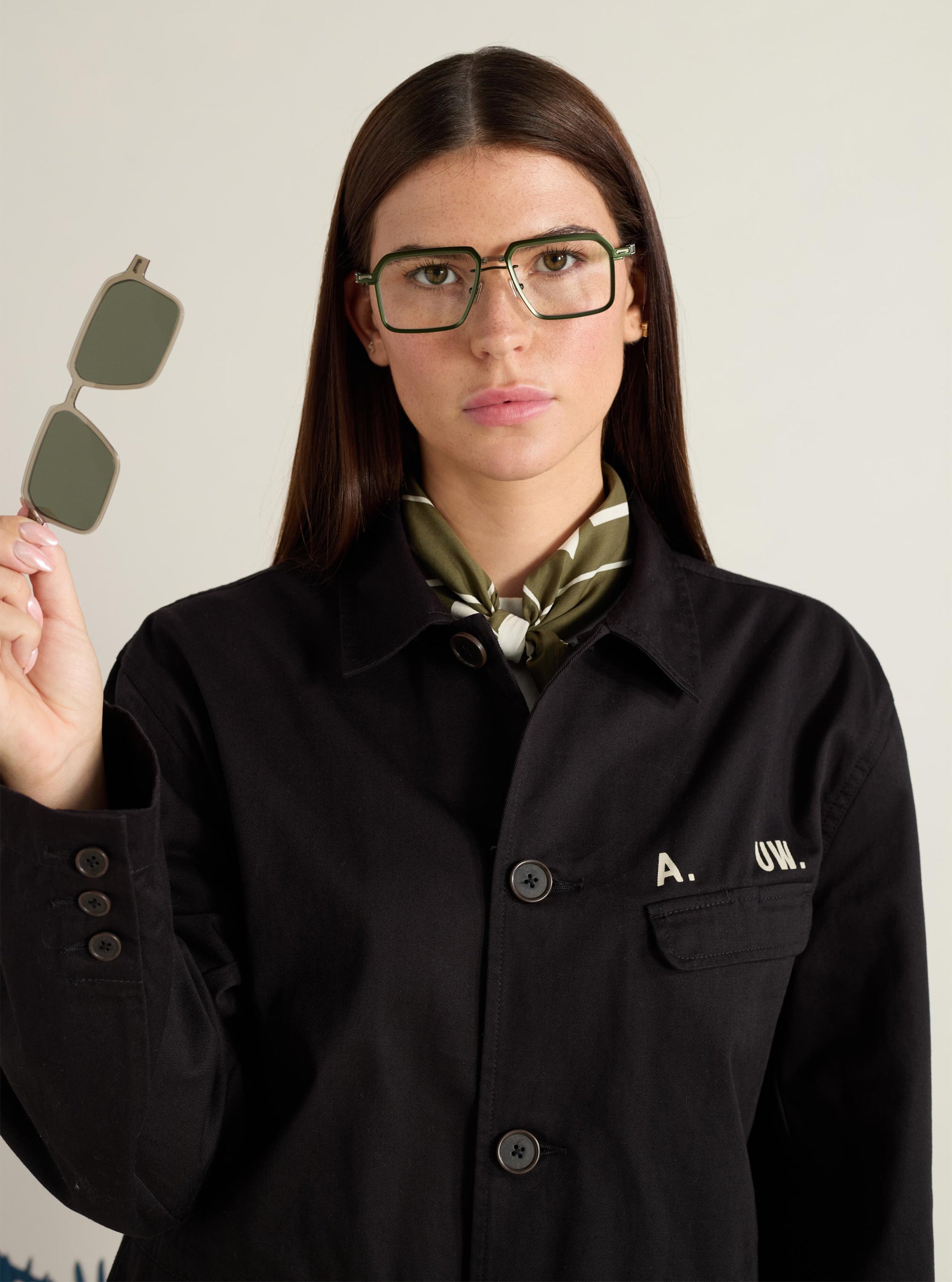 Model presents A. SOCIETY x Universal Works Olive/Green Kowloon Eyewear. She holds the olive clip-on lenses. The glasses have a green and gold metal frame. Studio shot on a neutral background.