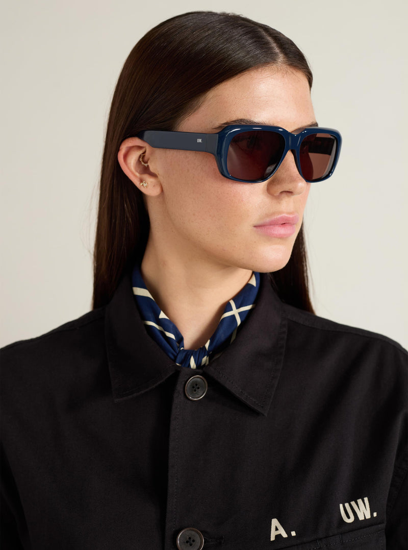 Model wearing A. SOCIETY x Universal Works Navy Aberdeen Sunglasses. The sunglasses have a thick navy frame and brown lenses. Shot in a studio with a neutral background.
