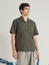 The Olive Tech Cotton S/S Dwight Overshirt is presented as a flat lay against a plain white background. The short-sleeved overshirt features a camp collar, a full button-down front, and four large patch pockets on the front. Two vertical rows of pleated details run down each side of the central closure. The garment is made from a smooth, olive green technical cotton fabric.