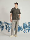 The Olive Tech Cotton S/S Dwight Overshirt is shown as a flat lay against a white background. The short-sleeved overshirt features two rows of vertical pleats sewn down the front, a camp collar, a central button closure, and four front patch pockets. The fabric is a smooth olive green cotton with a technical appearance.