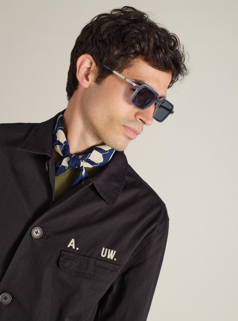 A model wears A. SOCIETY x Universal Works Silver/Grey Kowloon Eyewear. The eyewear has a grey frame and dark lenses. Shot in a studio with a neutral background.