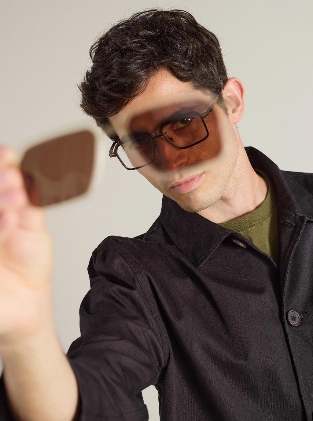 A model holds A. SOCIETY x Universal Works Kowloon Eyewear in brown/honey. The glasses have a square frame. Shot in a studio with a neutral background.