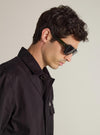 A model showcases A. SOCIETY x Universal Works Green Aberdeen Sunglasses. The sunglasses have a green frame. Shot in a studio with a plain background.
