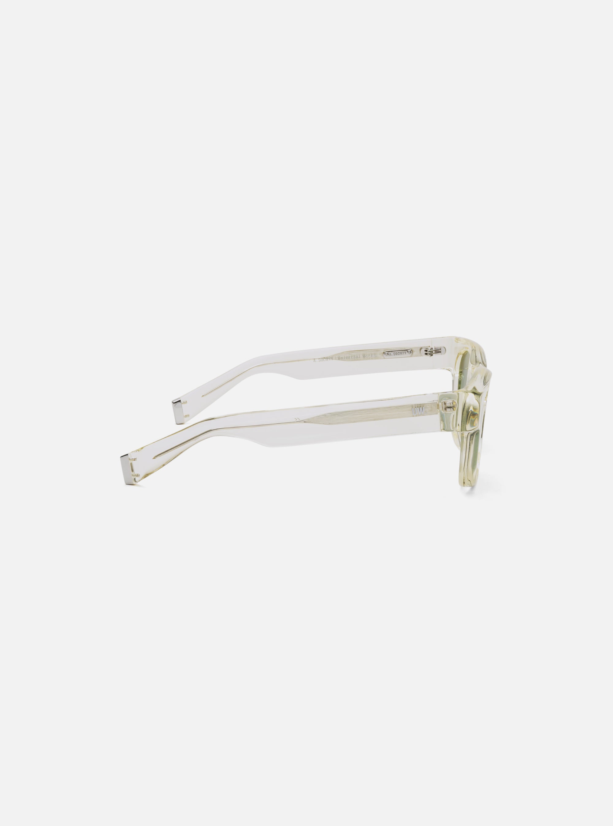 Angled view of A. SOCIETY x Universal Works Honey Stanley Sunglasses on a white surface. The frames are a translucent honey color with silver accents.