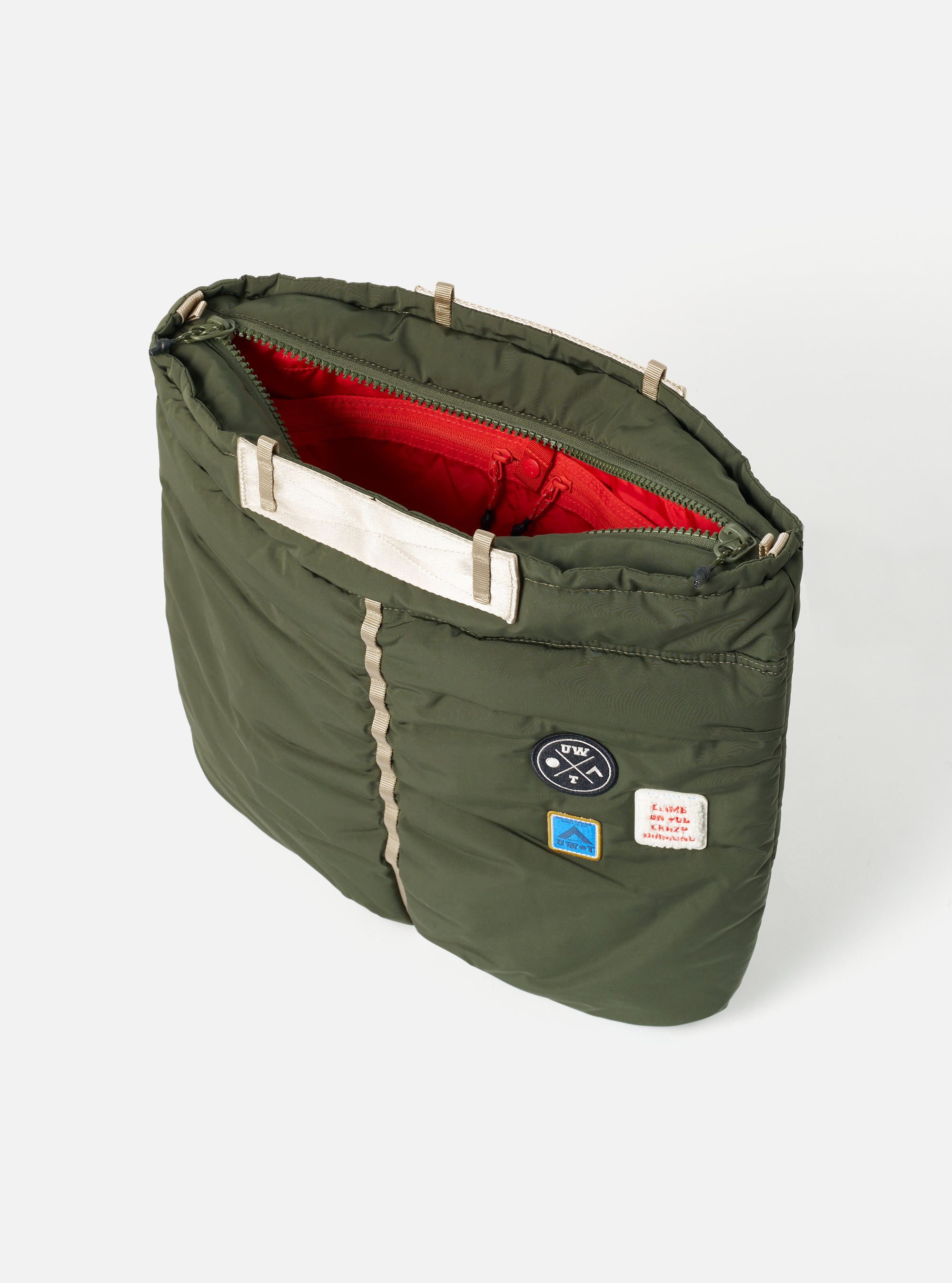 The Topologie x Universal Works Rifle Green UW Tech Helmet Bag on a white background. The bag is open, revealing a red interior. Patches adorn the exterior.