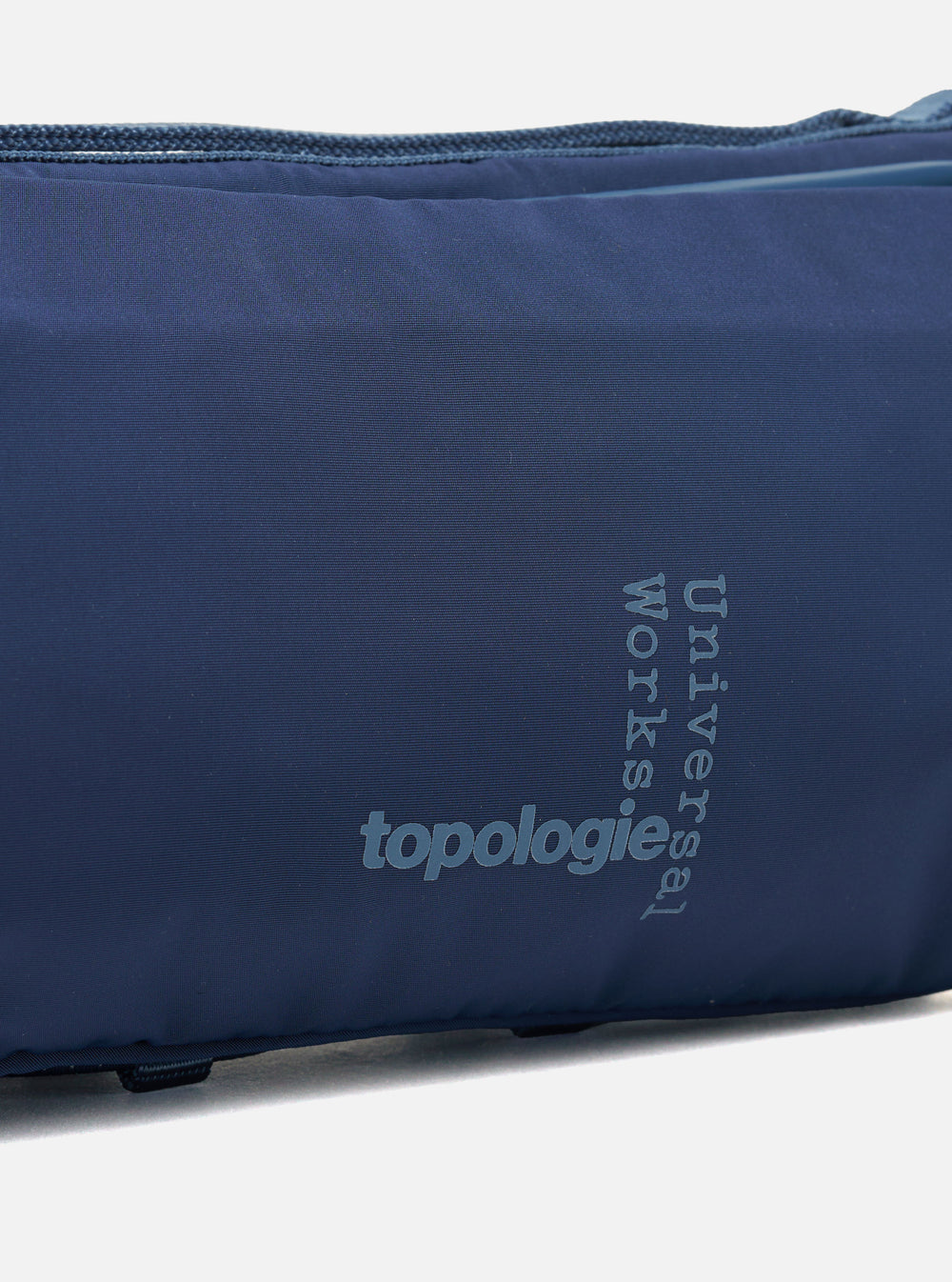 Close-up of the Topologie x Universal Works Insignia Blue UW Tech Bottle Sacoche Medium. The blue fabric bag features the logos of both brands in a lighter blue.