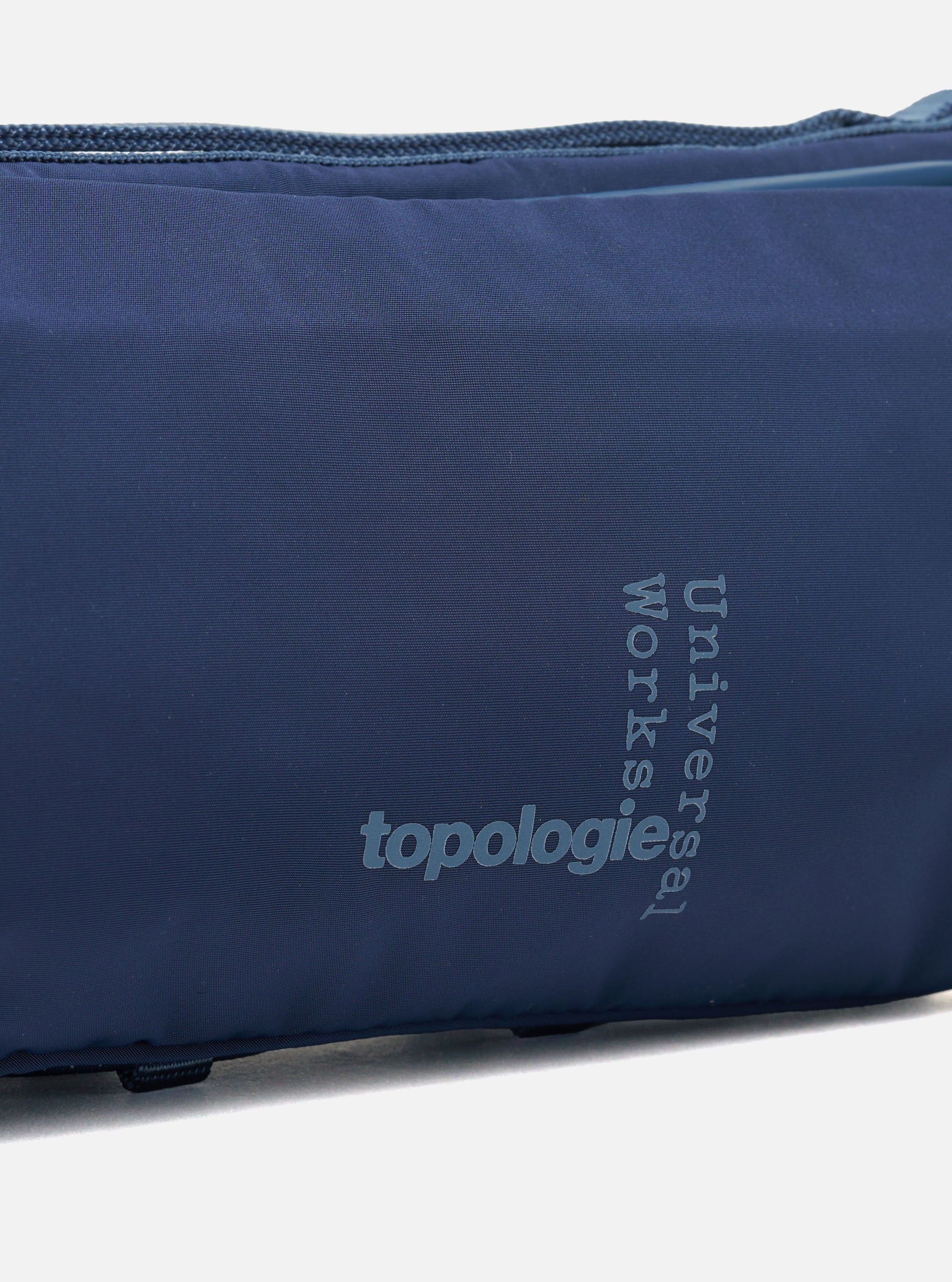Close-up of the Topologie x Universal Works Insignia Blue UW Tech Bottle Sacoche Medium. The blue fabric bag features the logos of both brands in a lighter blue.