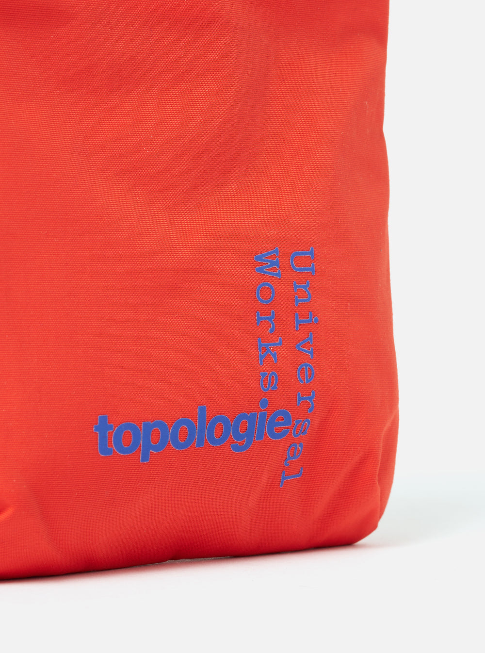 Close-up of the Topologie x Universal Works Red UW Tech Phone Sacoche. The red fabric bag features blue logos. Shot against a white background.