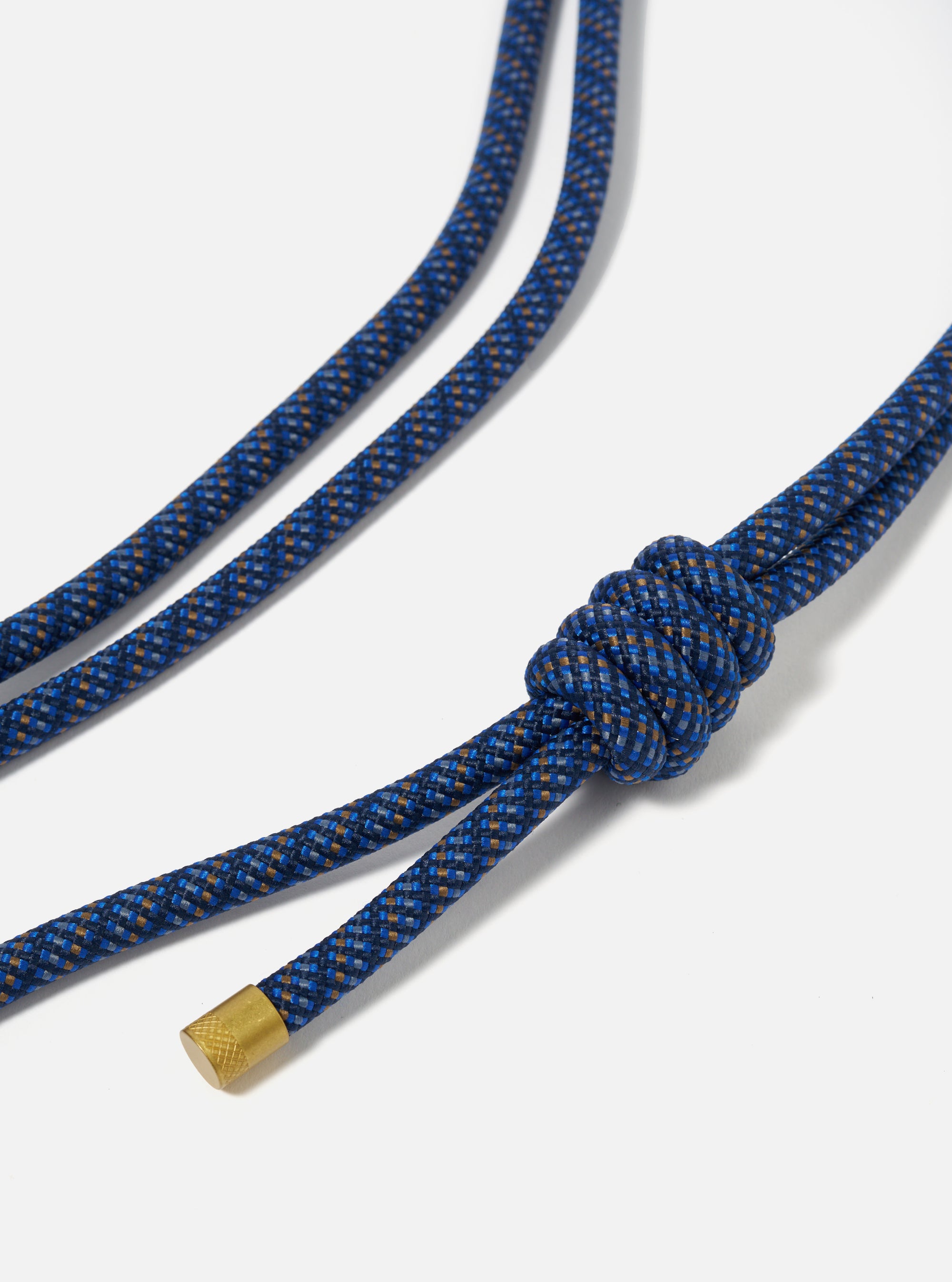 Close-up of the Topologie x Universal Works Navy Hitch UW Rope 8mm Rope Strap on a white surface. The navy and brown woven rope features a knot and gold metal end.