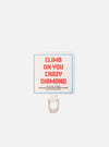 Close-up of the Topologie x Universal Works Off White UW Tech Phone Strap Adapter. Features an off-white fabric with red text and a blue border, paired with a silver metal loop on a white background.