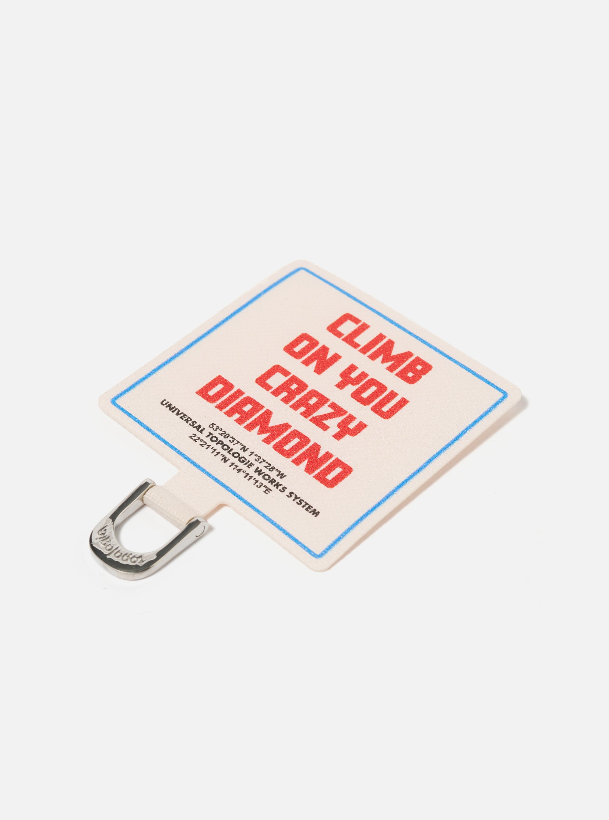 Close-up of the Topologie x Universal Works Off White UW Tech Phone Strap Adapter. It's off-white with red text and a blue border, featuring a silver metal clip. Shot on a clean white background.