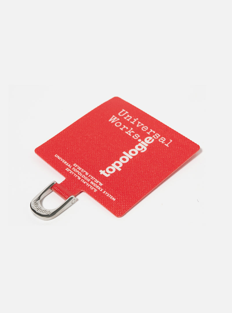 Close-up of the Topologie x Universal Works Red UW Tech Phone Strap Adapter on a white surface. The red fabric adapter features white text and a silver metal loop.