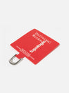 Close-up of the Topologie x Universal Works Red UW Tech Phone Strap Adapter on a white surface. The red fabric adapter features white text and a silver metal loop.