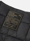 Close-up of the TAION x Universal Works Reversible Black Military Line Vest, showcasing the label with brand info on a quilted black fabric. Studio shot.