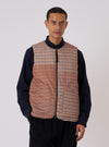 A model poses in the TAION x Universal Works Reversible Black Military Line Vest. The vest is brown plaid with a black zipper. He wears a navy shirt and black pants against a white backdrop.