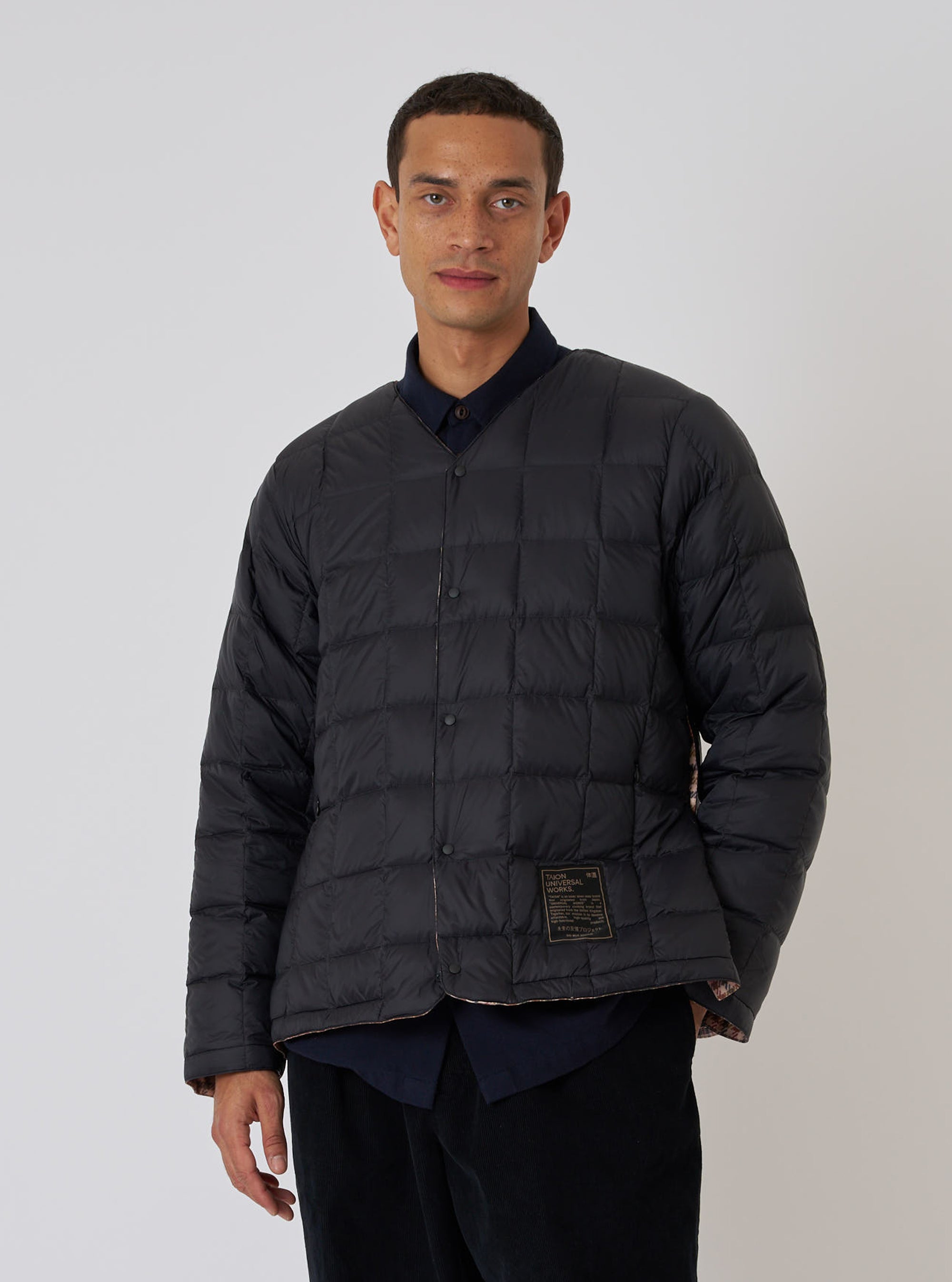 A model poses in a TAION x Universal Works Reversible Black Military Line Liner Jacket against a white background. The quilted jacket is buttoned and worn over a dark shirt.