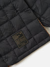 Close-up of the TAION x Universal Works Reversible Black Military Line Liner Jacket. Features black quilted material, a branded tag, and a glimpse of the reversible patterned lining.