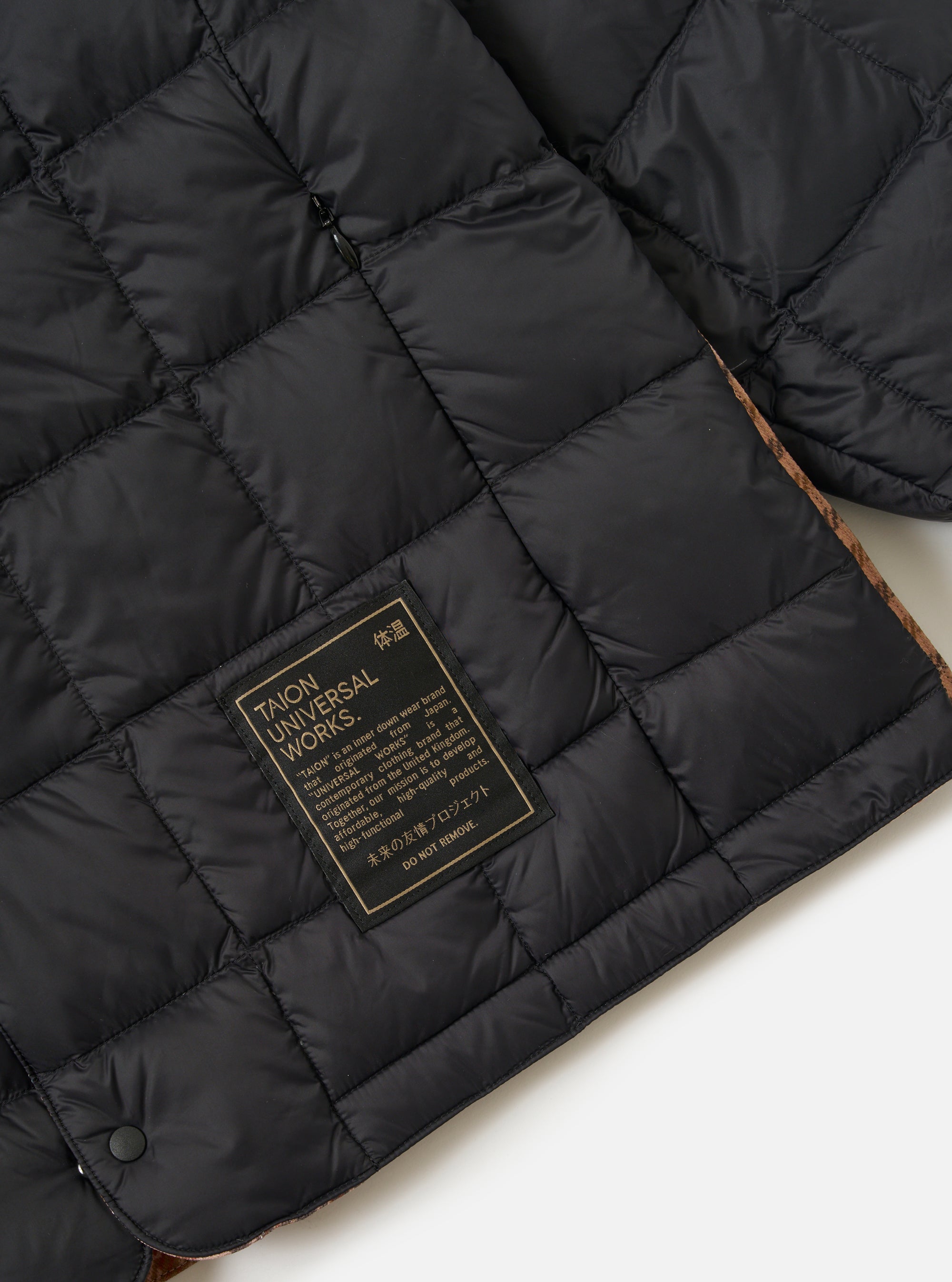 Close-up of the TAION x Universal Works Reversible Black Military Line Liner Jacket. Features black quilted material, a branded tag, and a glimpse of the reversible patterned lining.