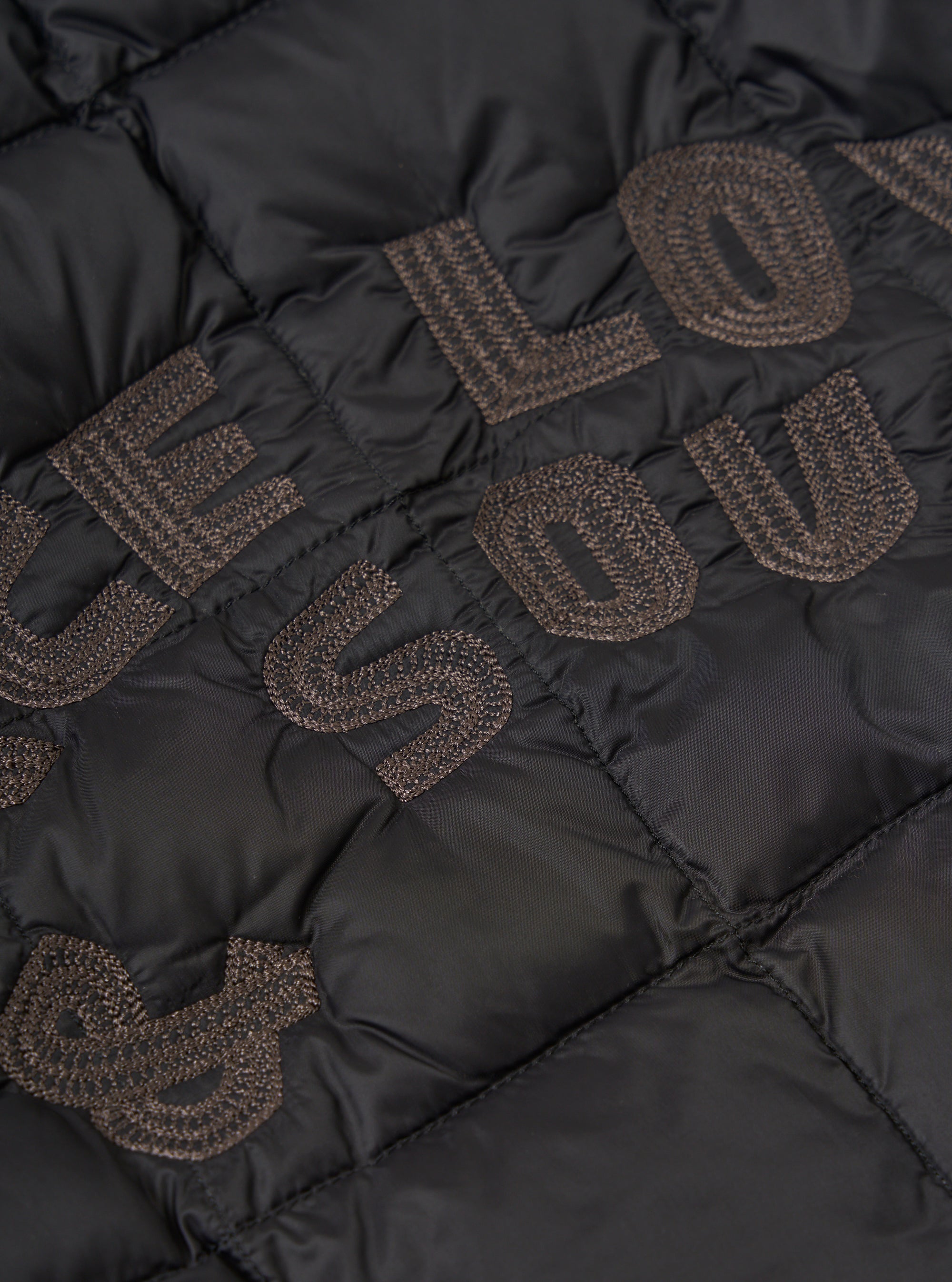 Close-up of the TAION x Universal Works Reversible Black Military Line Liner Jacket. Features black quilted fabric with embroidered brown lettering. 