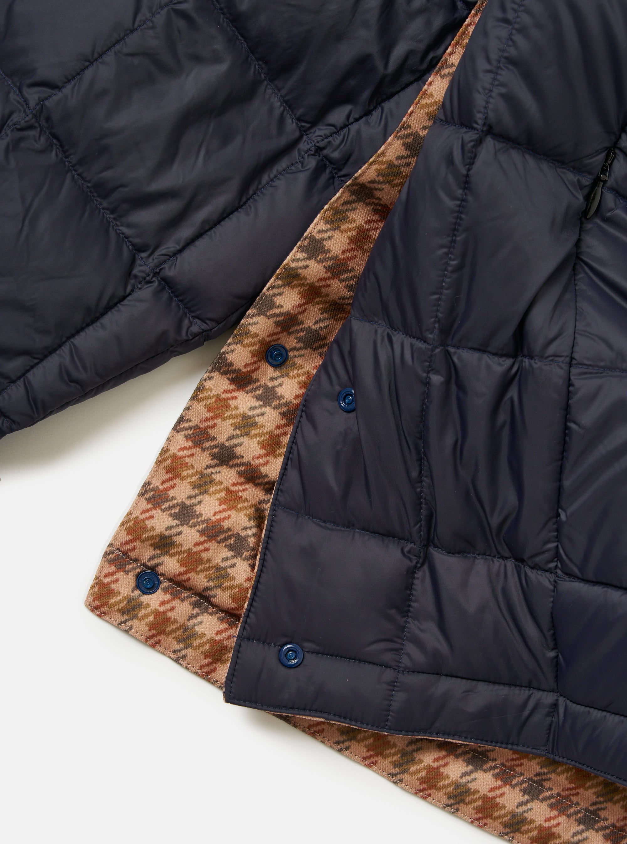 Close-up of the TAION x Universal Works Reversible Dark Navy Military Line Liner Jacket on a white surface. Shows the dark navy quilted material and the brown houndstooth reversible side.