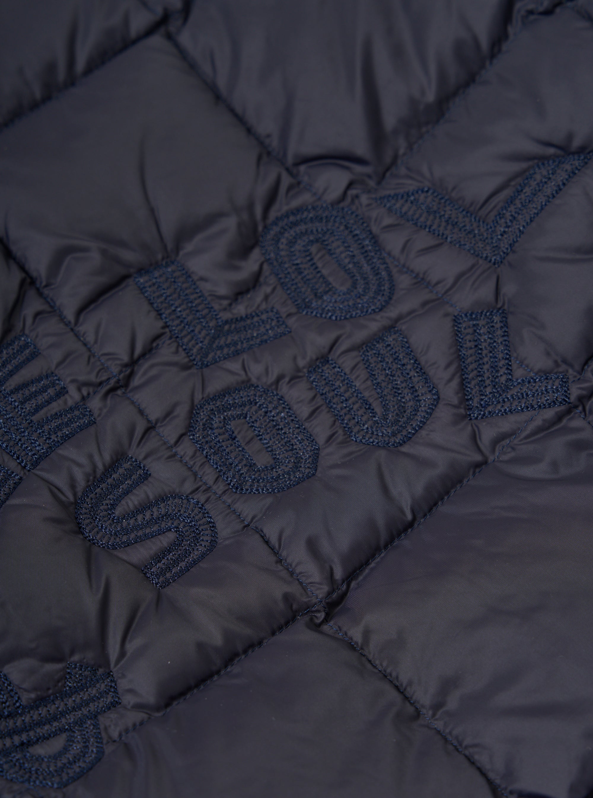 Close-up of the TAION x Universal Works Reversible Dark Navy Military Line Liner Jacket, showcasing the quilted texture and embroidered lettering on the dark navy material.