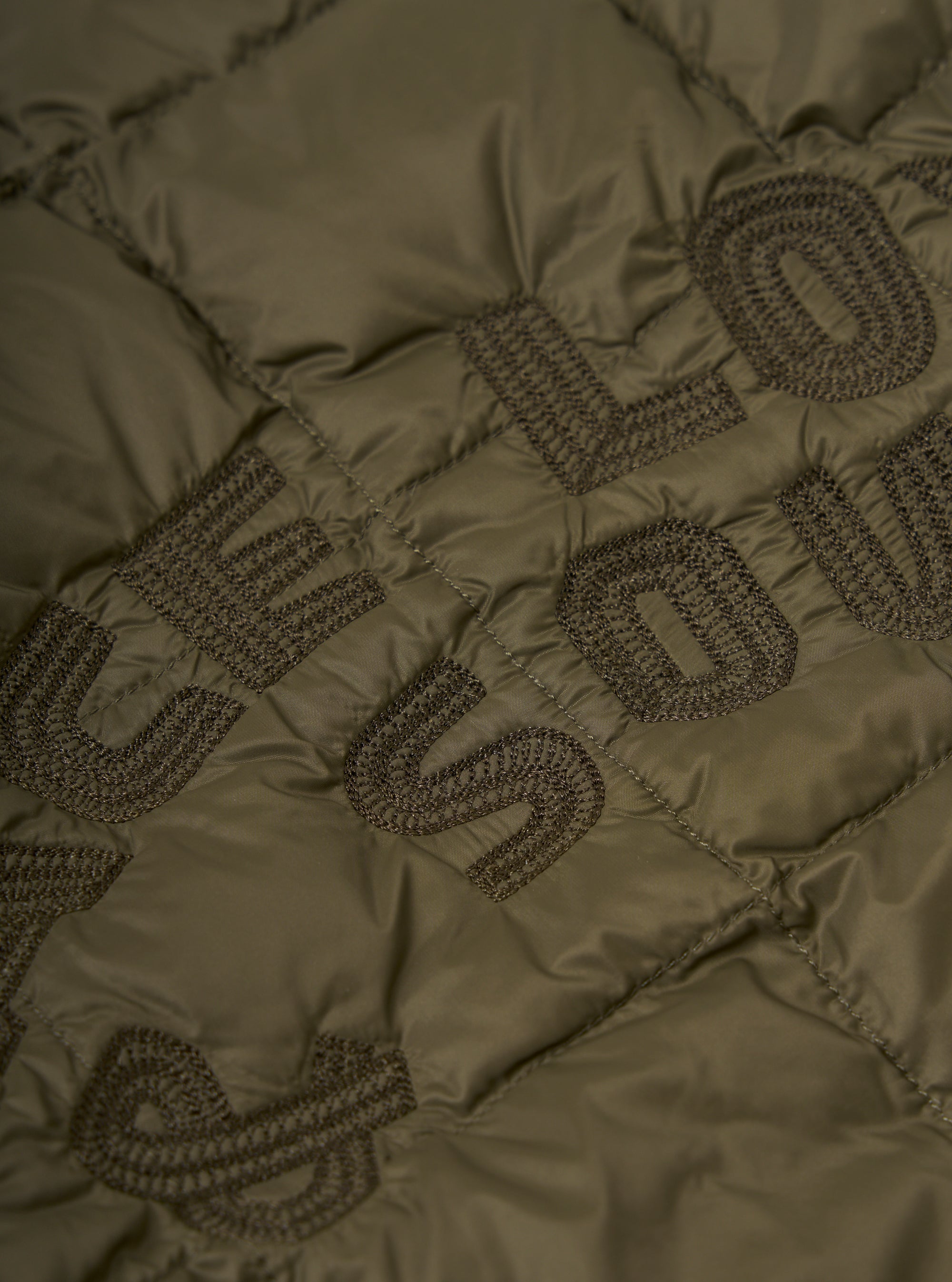 Close-up of the TAION x Universal Works Reversible Dark Olive Military Line Liner Jacket, showcasing the quilted texture and embroidered logo detail on the olive green fabric.