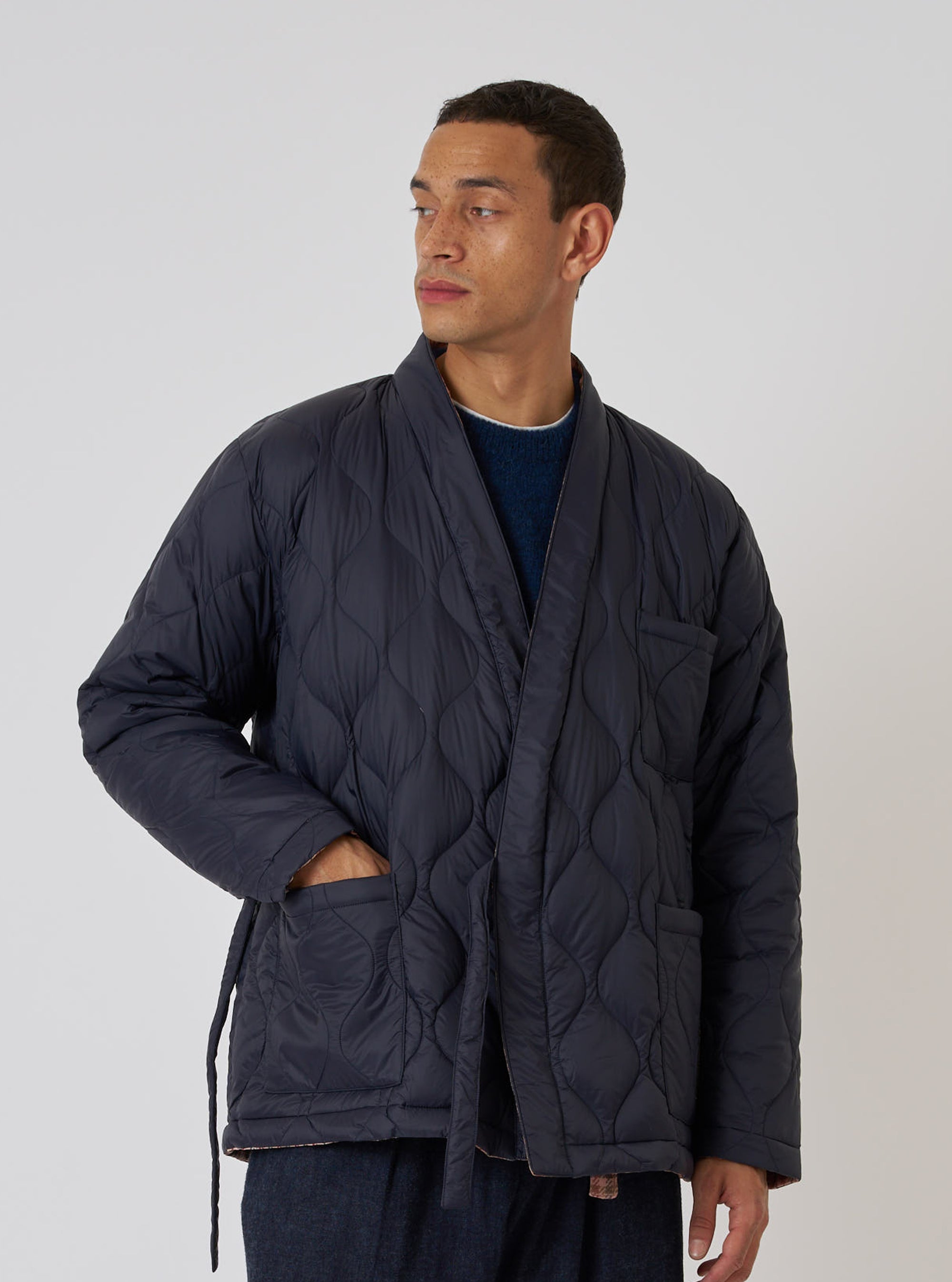 A model wears the TAION x Universal Works Reversible Dark Navy Military Line Kyoto Work Jacket. The quilted jacket has a tie waist and is set against a plain background.