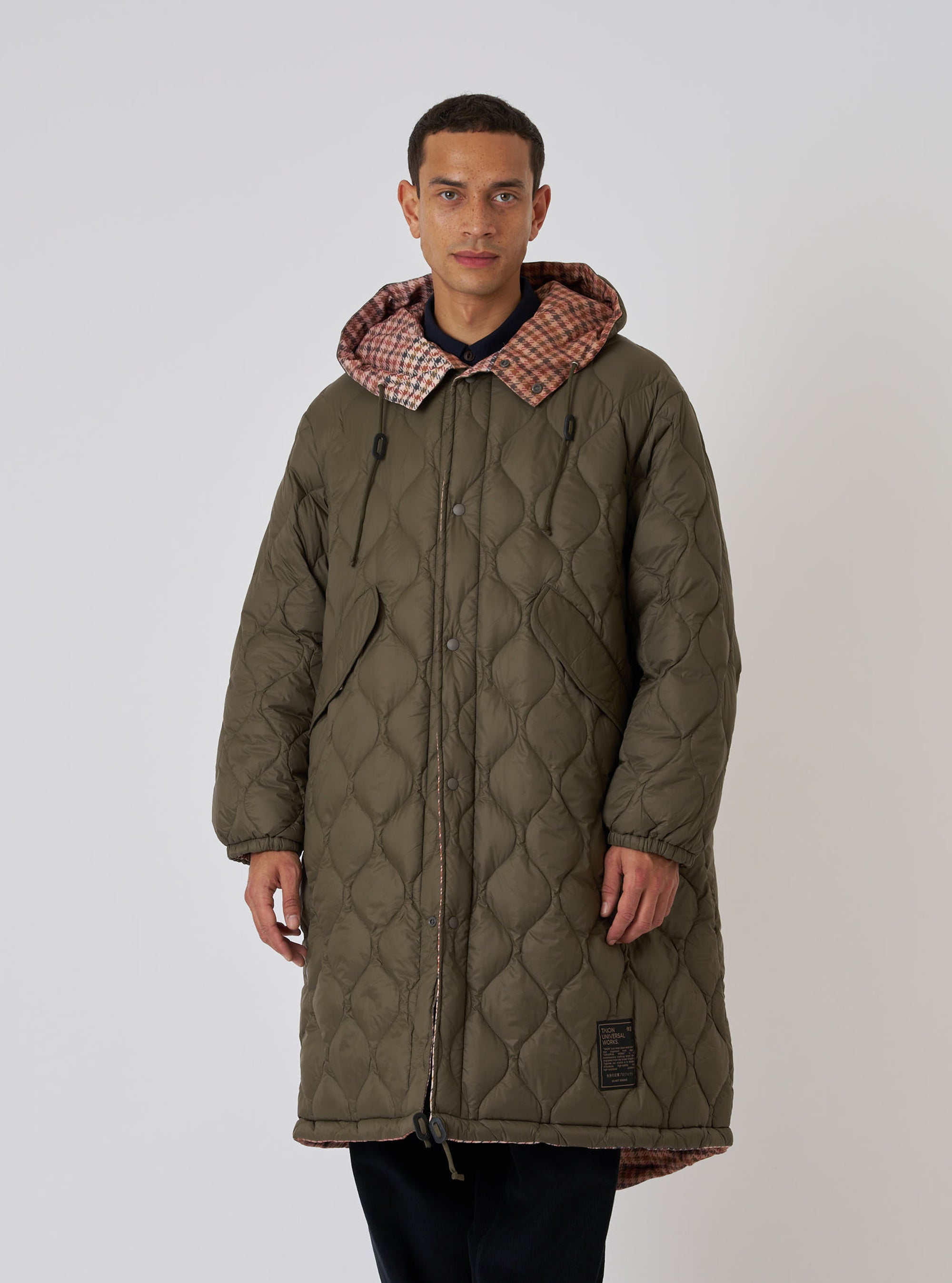A model in a TAION x Universal Works Reversible Dark Olive Military Line Parka. The quilted parka has a plaid hood lining. Shot on a white background.