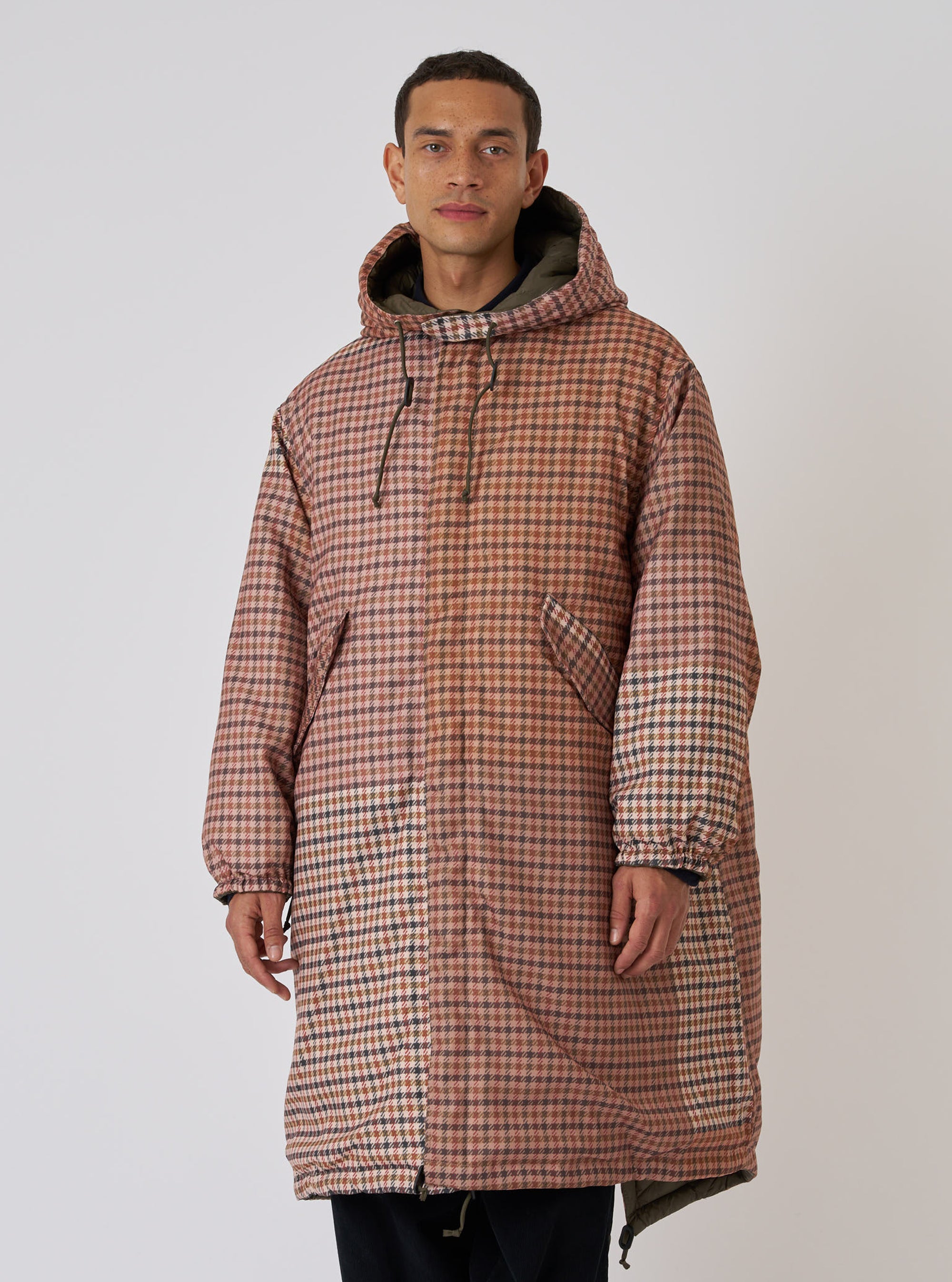 A model on a white background wears the TAION x Universal Works Reversible Dark Olive Military Line Parka. The parka has a brown and tan houndstooth pattern and a relaxed fit.
