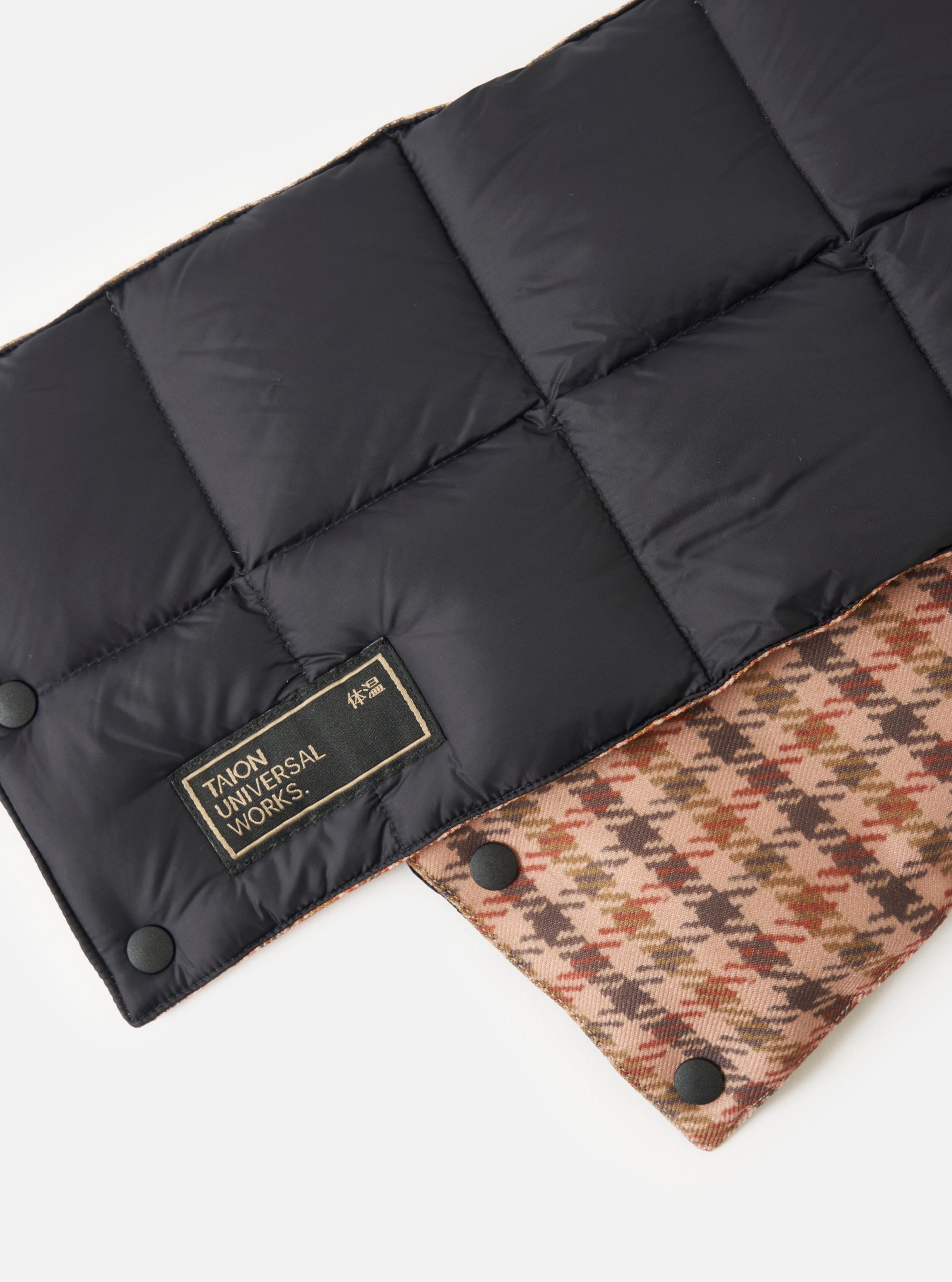Close-up of the TAION x Universal Works Reversible Black Military Line Neck Warmer on a white surface. One side is black quilted nylon, the other is a brown and tan houndstooth pattern.