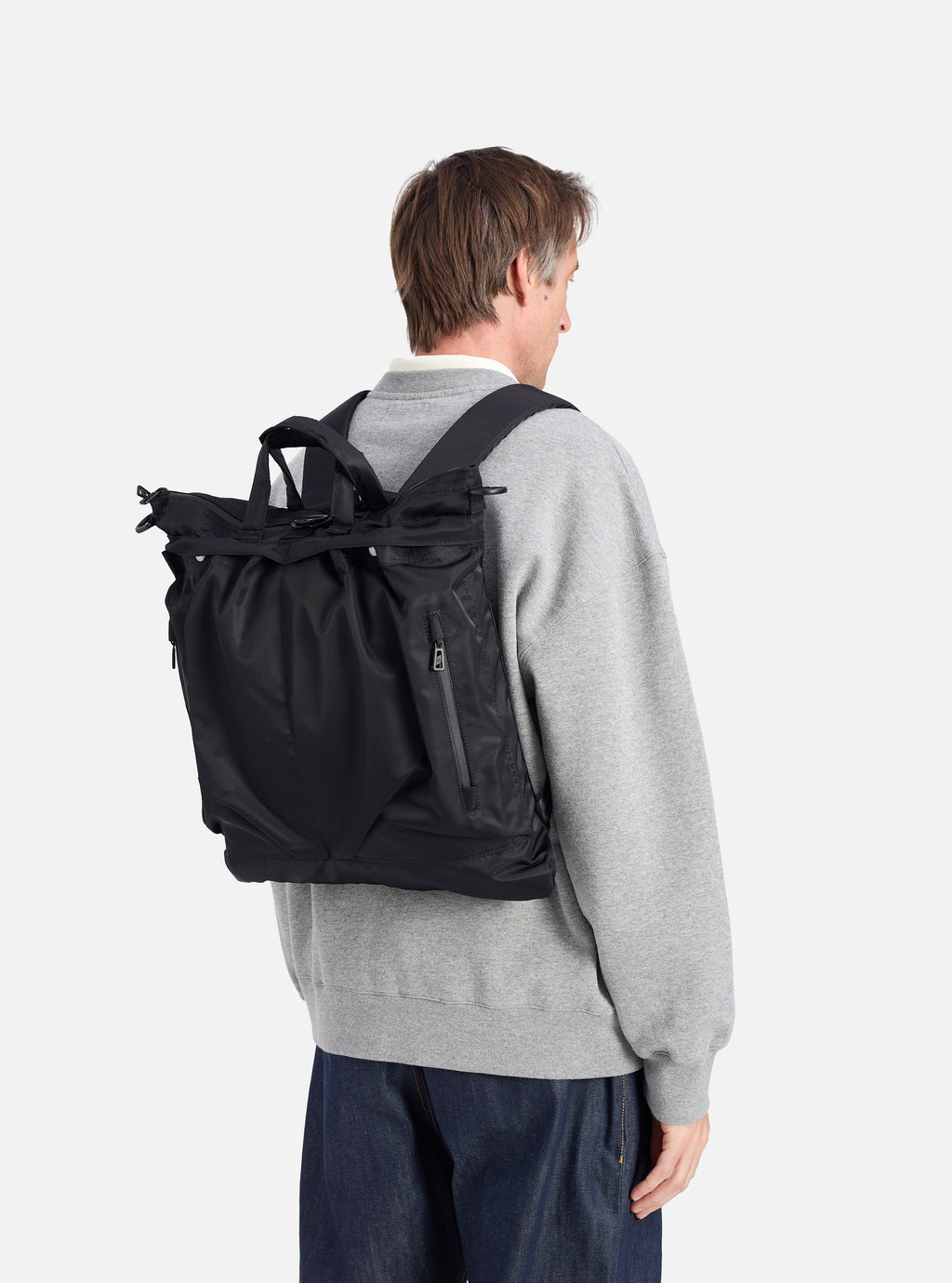 Back view of a model wearing the F/CE.® Black Recycled Twill 3-Way Helmet Bag as a backpack. The bag is black and made of twill. The background is a clean white studio.