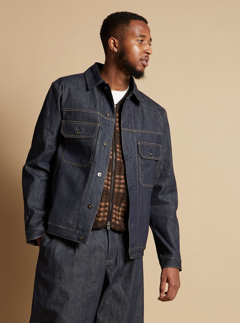 A model poses in an Indigo 13oz Selvedge Denim Trucker Jacket. The dark wash denim jacket is styled with a brown vest. Studio shot on a neutral background.