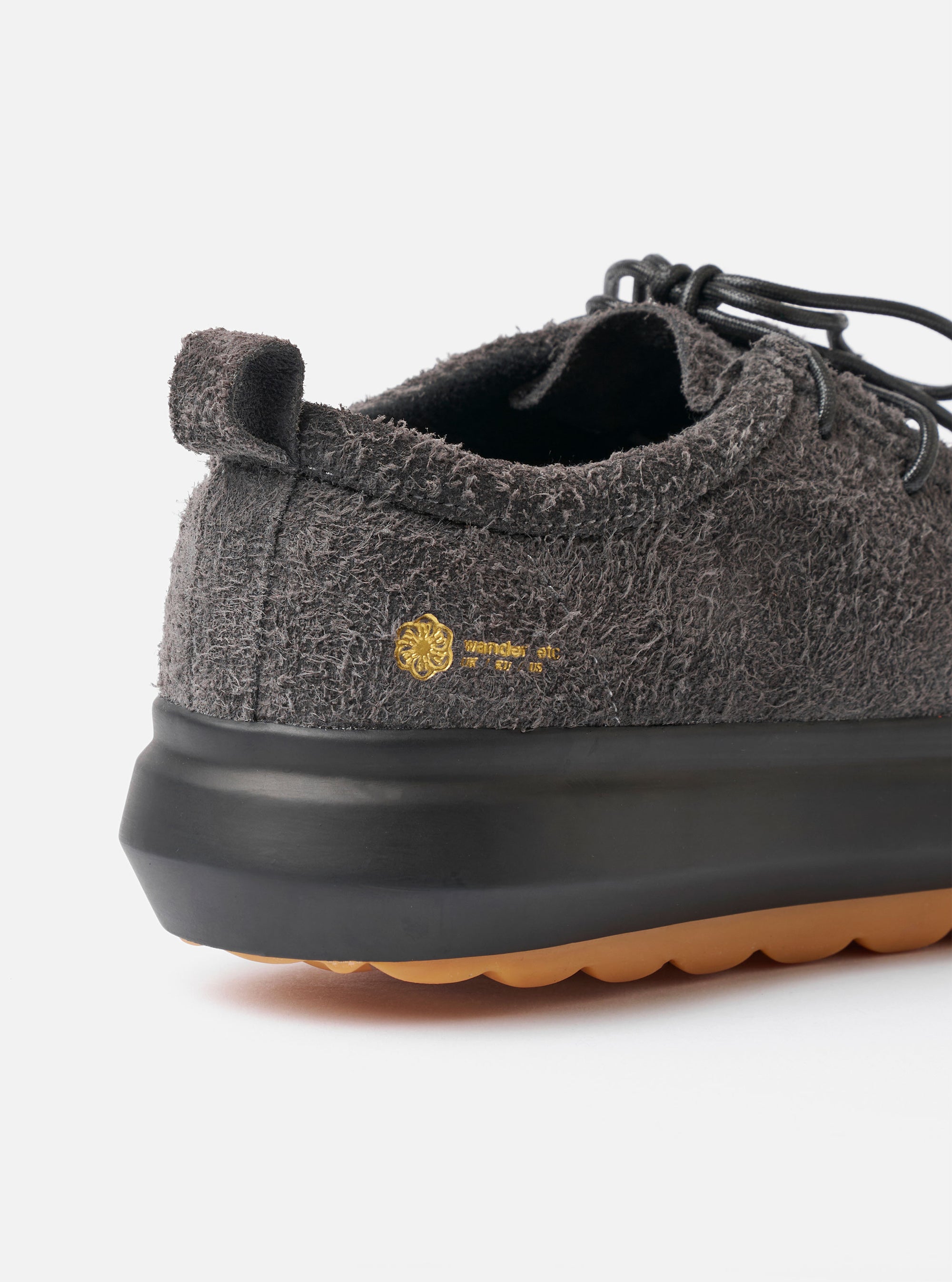 The Wander etc. FD/08 Low in Black Asphalt/Gum Hairy Suede is shown from a rear angle as a product shot against a plain white background. The image highlights the textured black hairy suede upper and the prominent chunky gum-colored rubber sole. The low-cut design features a vertical pull loop at the heel and tonal black stitching.