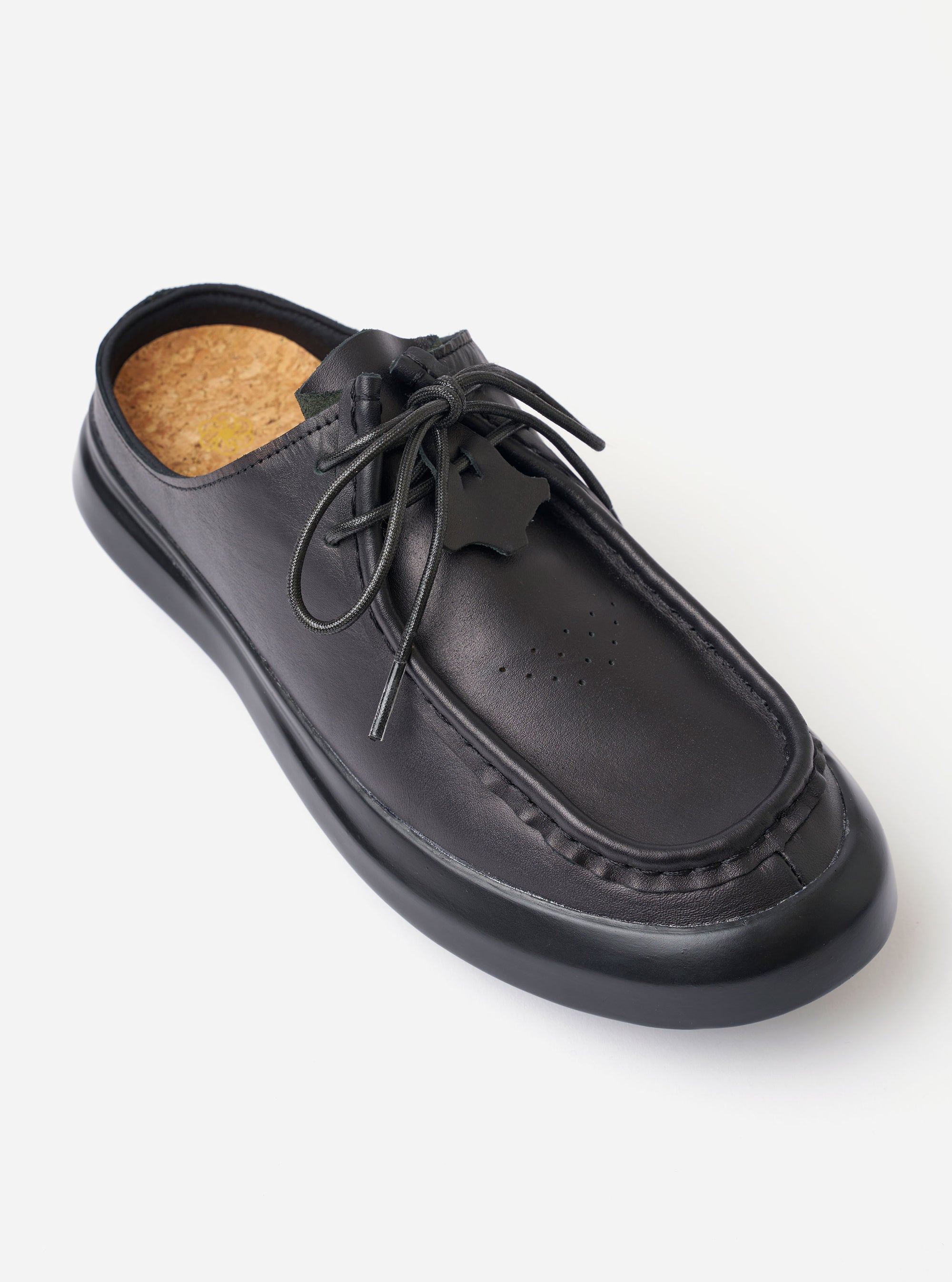 The Wander etc. Laze in Black Nappa Leather is shown from a rear three-quarter angle as a flat lay against a white background. The image highlights the backless mule design, the smooth black nappa leather upper, and the thick, chunky black outsole. The interior features a tan cork footbed with a printed logo.