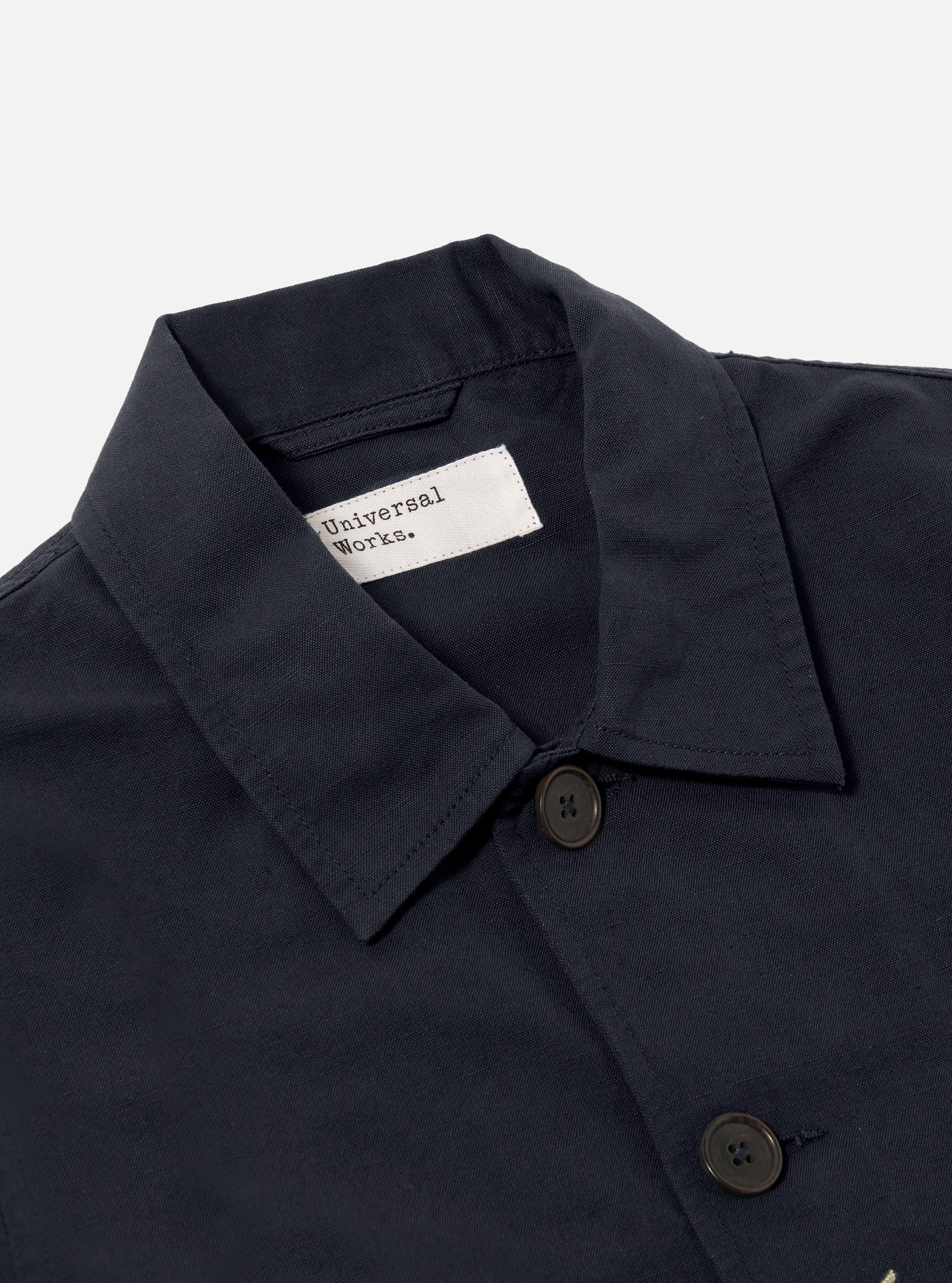 Close-up of the Navy Linen Slub Weave Bakers Jacket. The jacket is navy with brown buttons and a visible brand label. Shot on a plain white background.