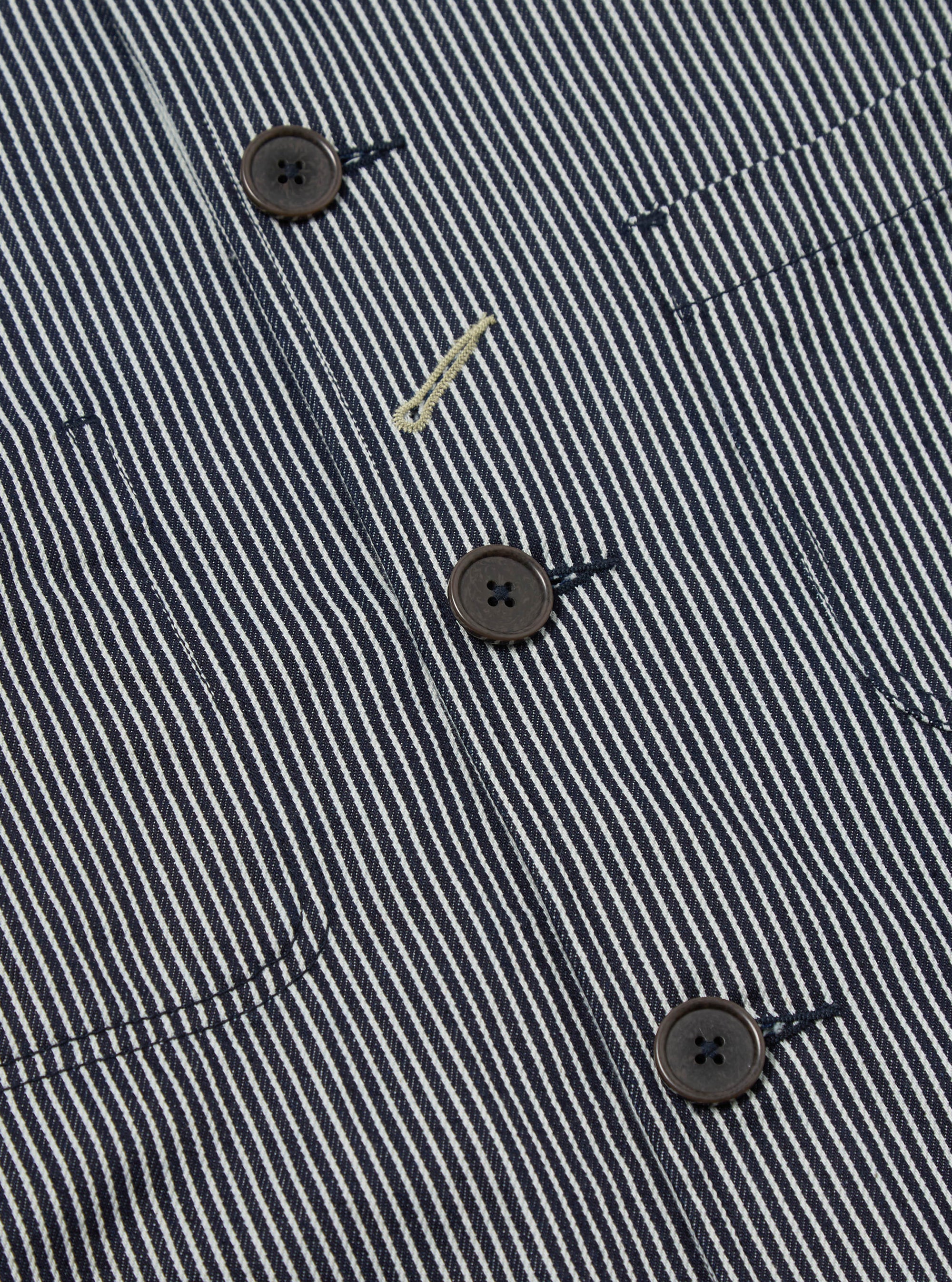 Close-up of the Indigo Hickory Stripe Denim Original Bakers Jacket. Features include brown buttons, a striped pattern, and denim material. Shot on a flat surface.