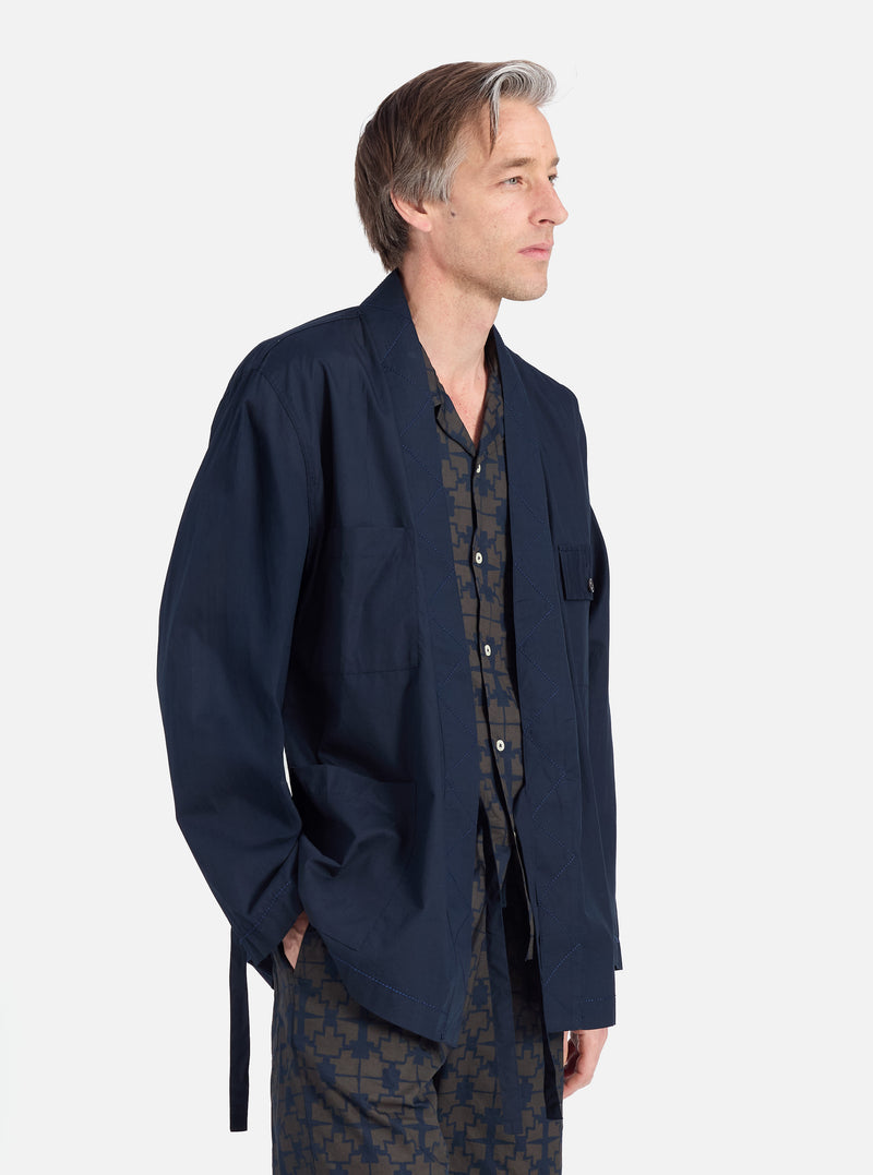 A man in profile on a white background wears the Navy Broadcloth Osaka Work Jacket. The jacket is broadcloth with a tie. He also wears a patterned shirt and pants.
