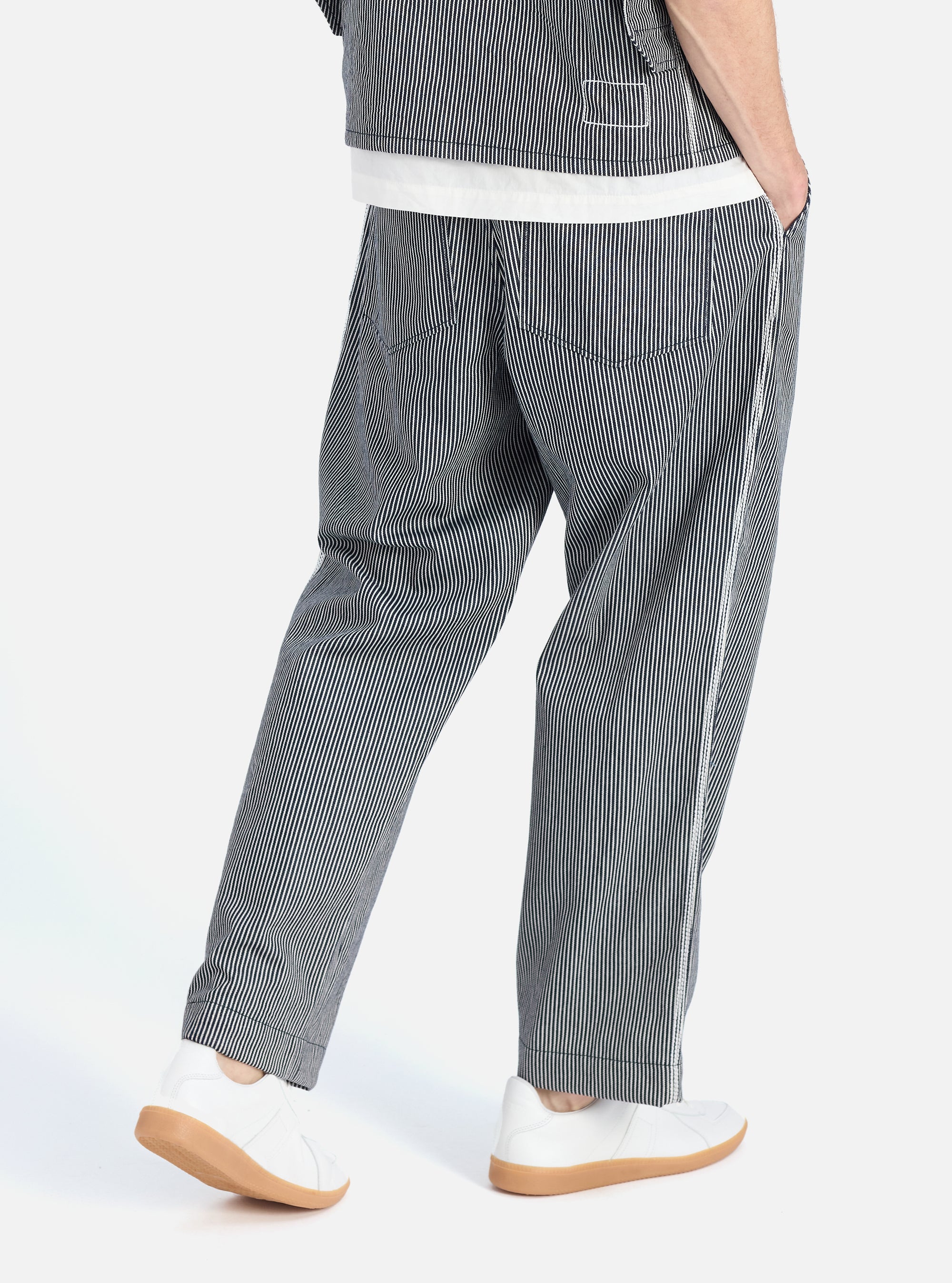 Model wearing Indigo Hickory Stripe Denim Duke Pant in studio. The relaxed-fit pants are blue and white striped denim. Worn with white sneakers. 