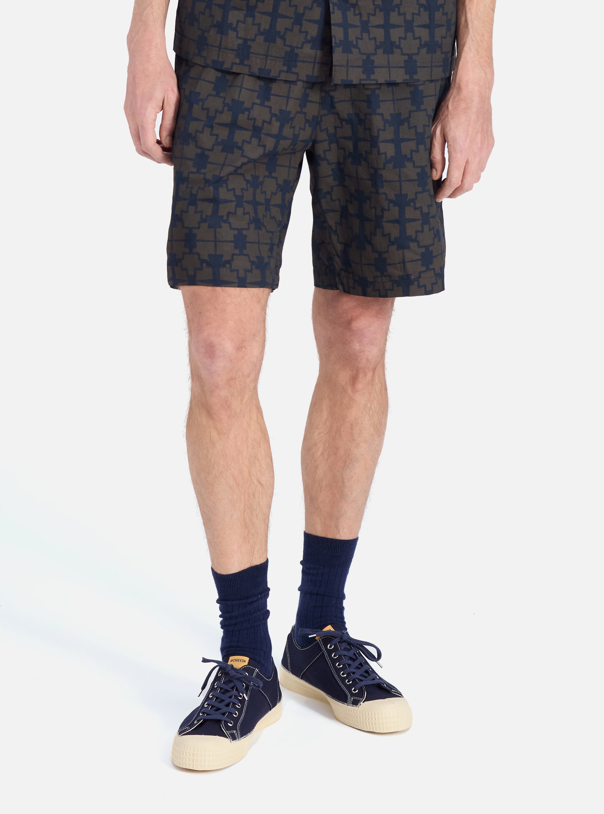 A man stands on a white background wearing the Navy Mitla Print Cotton Simple Short. The shorts have a navy and brown pattern. He also wears navy socks and sneakers.