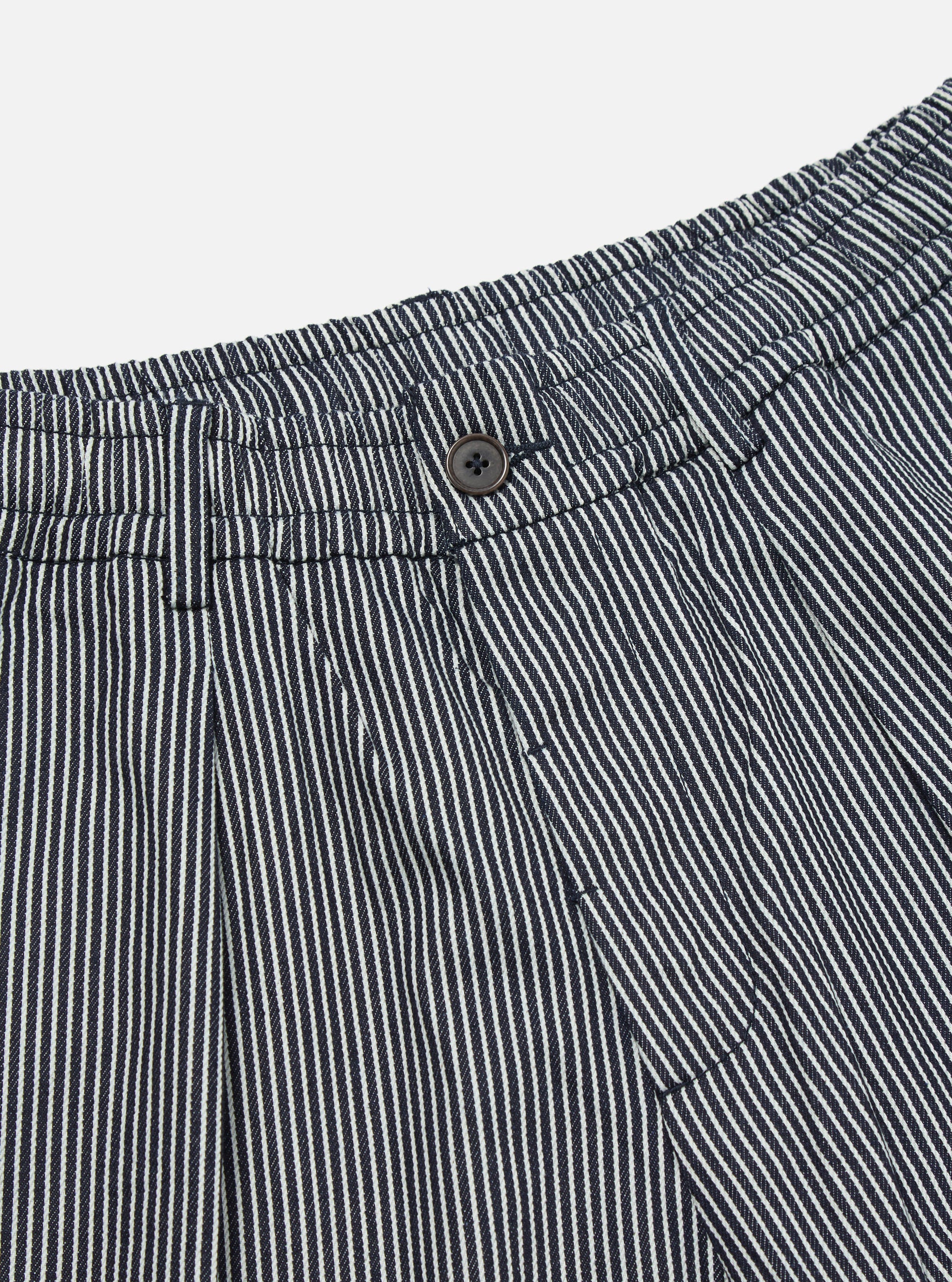 Close-up of the Indigo Hickory Stripe Denim Pleated Track Short. The denim is striped with white and indigo. Features a button closure and pleated design.