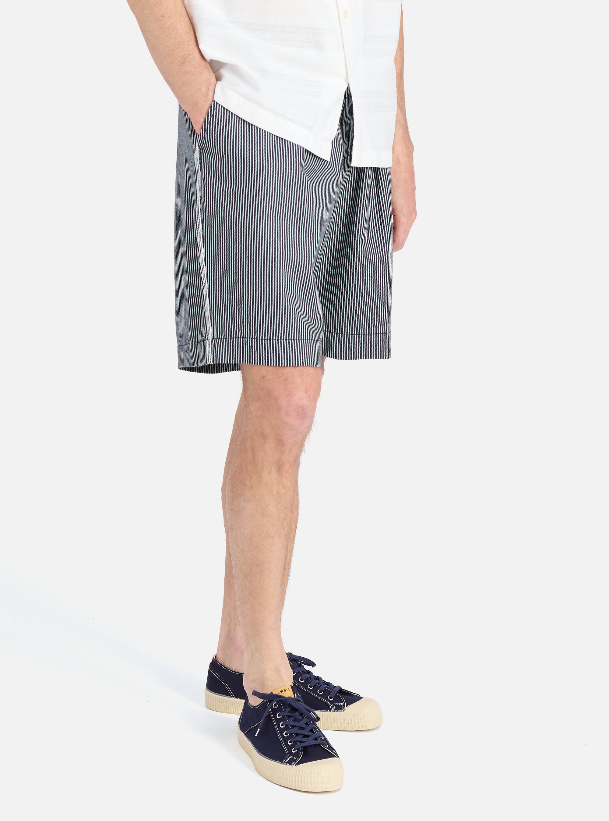 A model wears Indigo Hickory Stripe Denim Pleated Track Short, paired with a white shirt and blue sneakers, against a clean white backdrop.