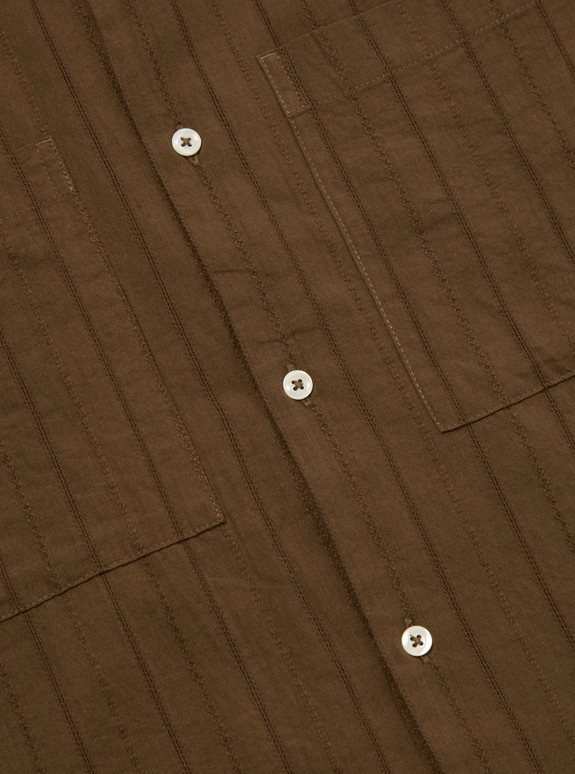 Close-up of the Brown Dobby Self Stripe Camper Shirt. The brown dobby fabric has self stripes. White buttons are visible. Two chest pockets. Shot on a flat surface.