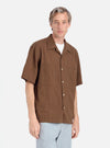 A man stands against a white backdrop, modeling the Brown Dobby Self Stripe Camper Shirt. The shirt has a relaxed fit and short sleeves. He is wearing light blue pants.