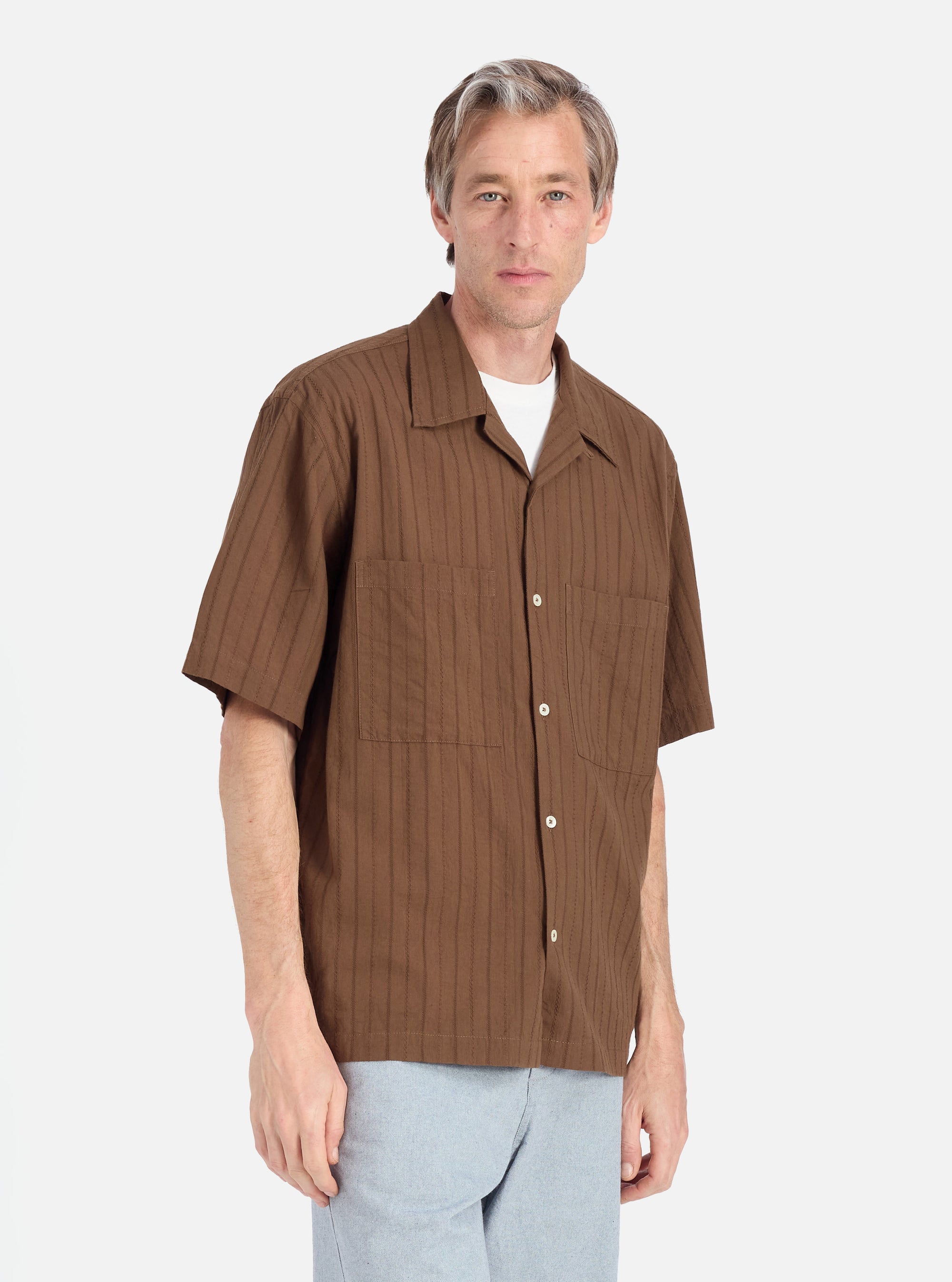 A man stands against a white backdrop, modeling the Brown Dobby Self Stripe Camper Shirt. The shirt has a relaxed fit and short sleeves. He is wearing light blue pants.