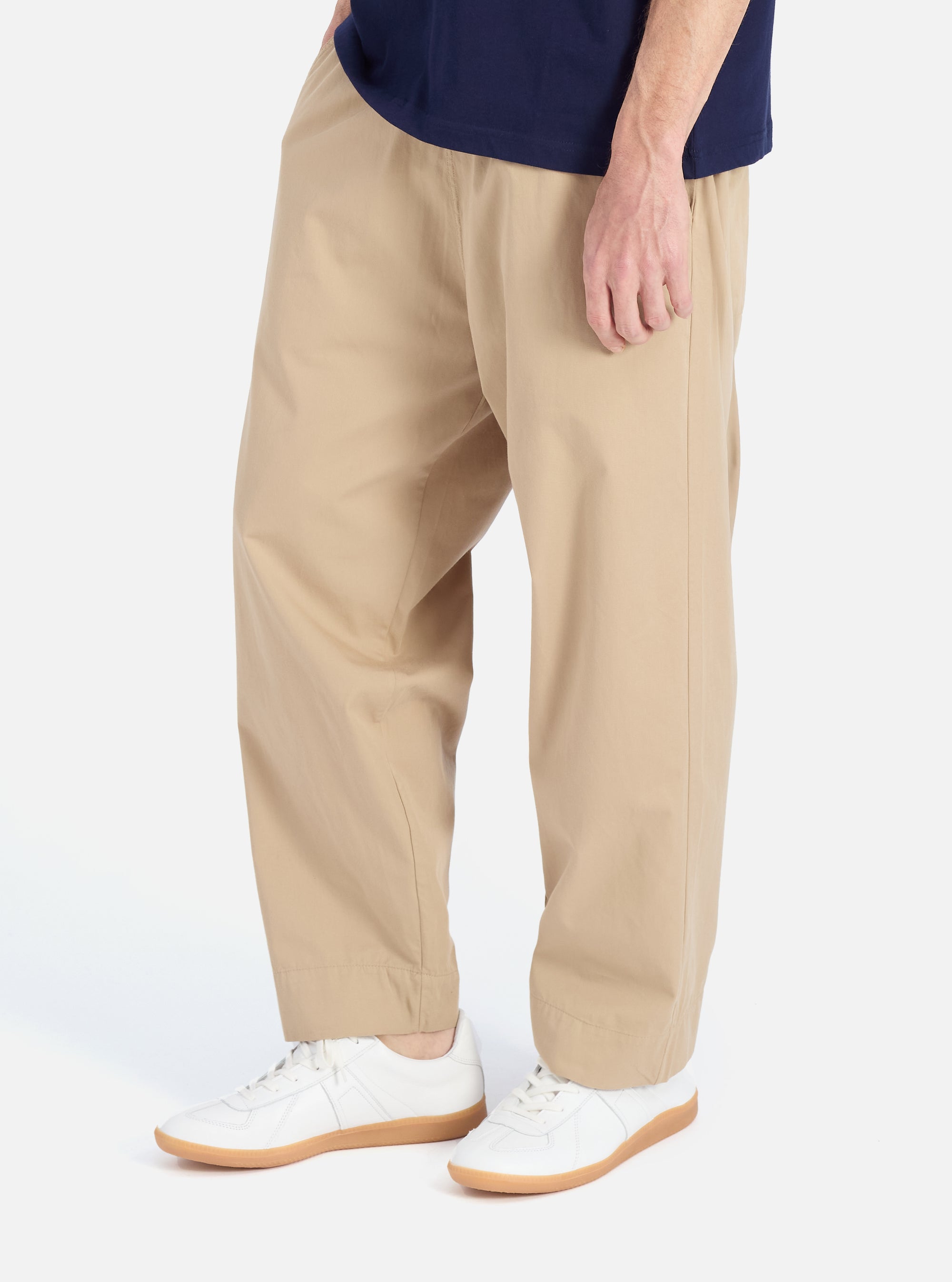A man models the Sand Military Cotton Judo Pant in a studio setting. The pants are a relaxed fit and paired with white sneakers. The background is a clean, bright white.